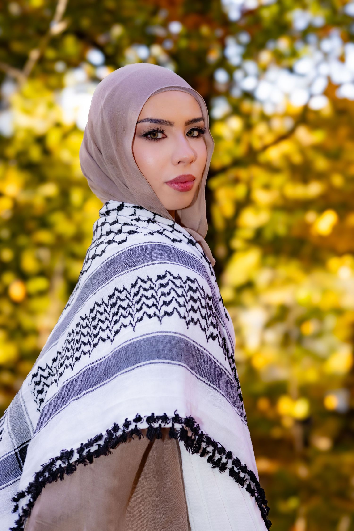 A person wearing a light gray hijab and black and white striped wrap poses against autumn foliage in soft lighting.