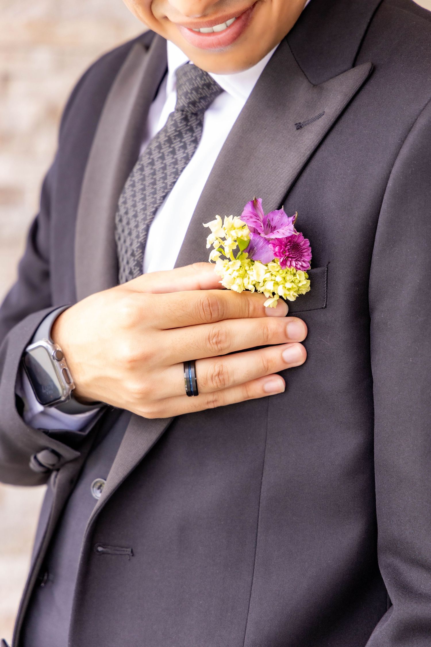 Vibrant purple and yellow boutonniere pinned to dark suit jacket with wedding ring visible.