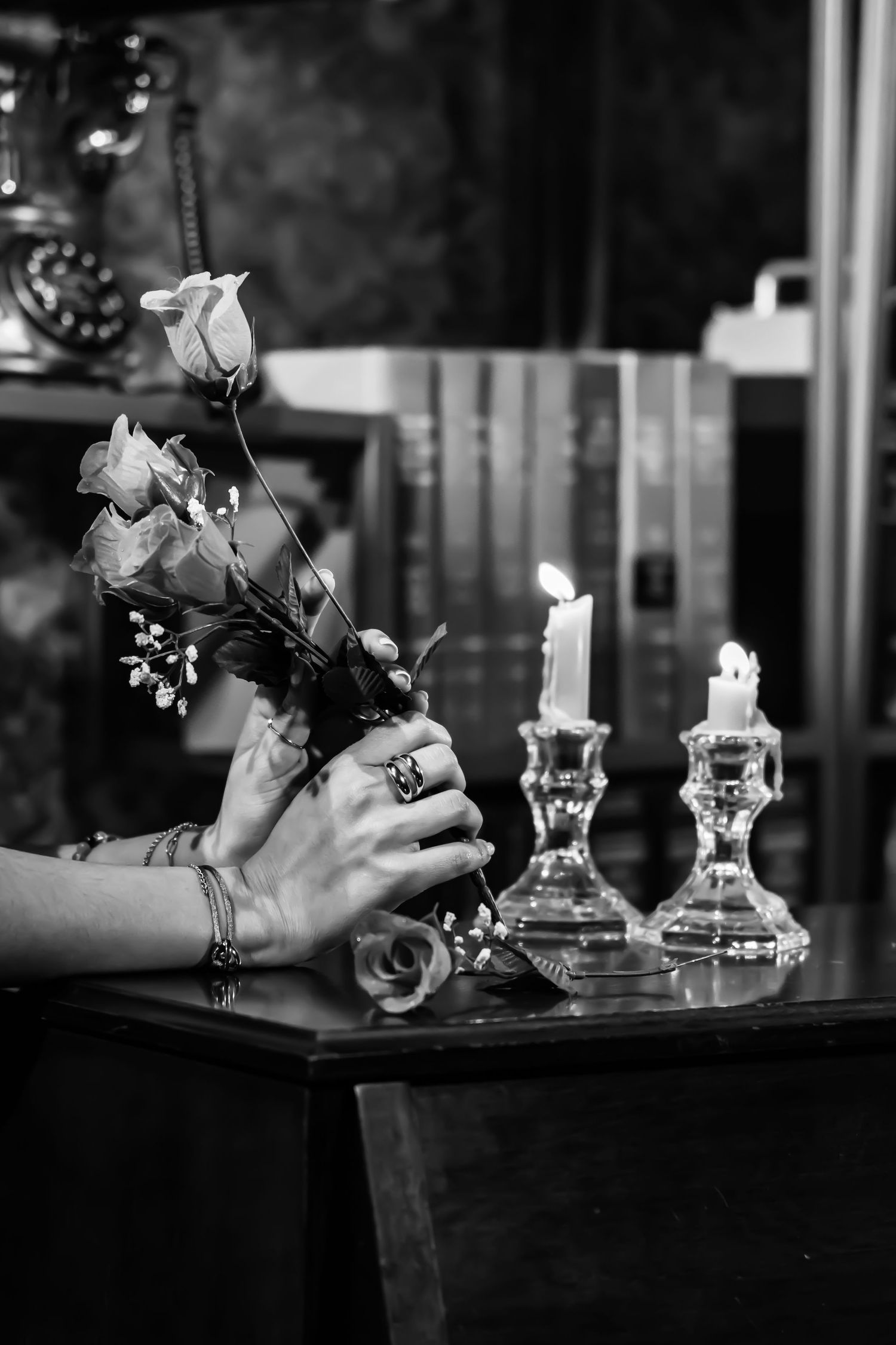Hands adorned with jewelry arrange white roses in a vase beside crystal candlesticks in moody black and white photography.