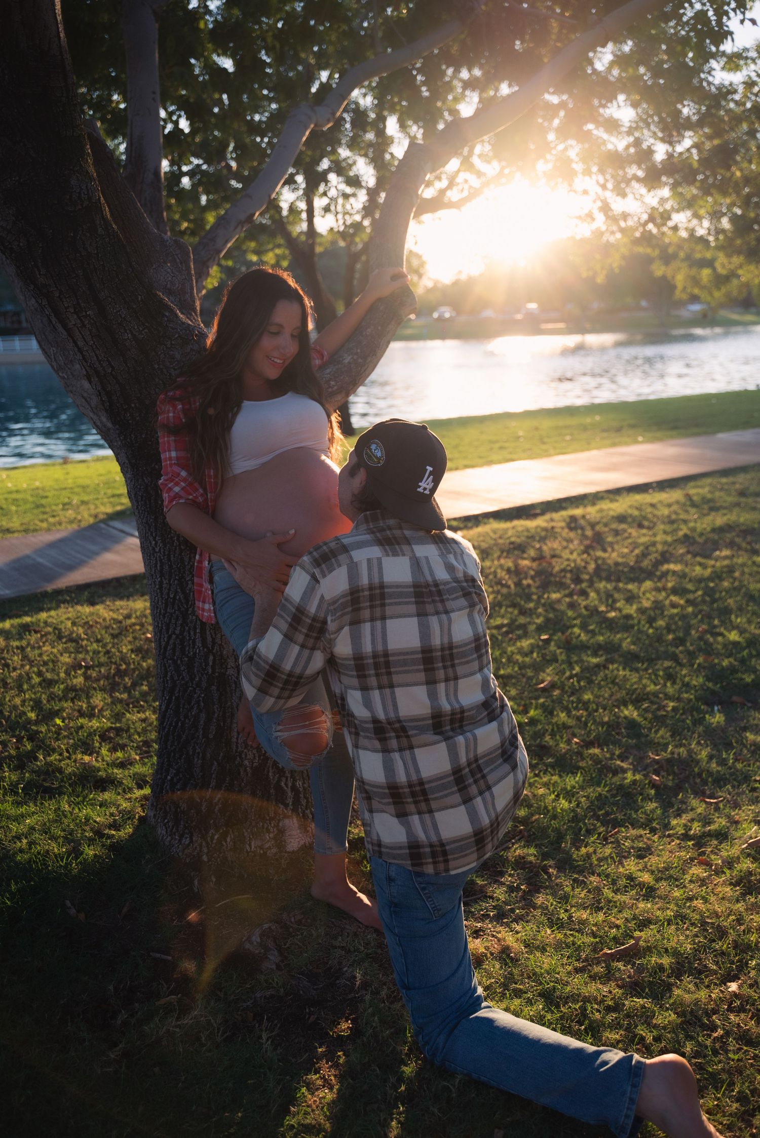 A couple embraces against a tree during golden hour sunset in a grassy park setting.