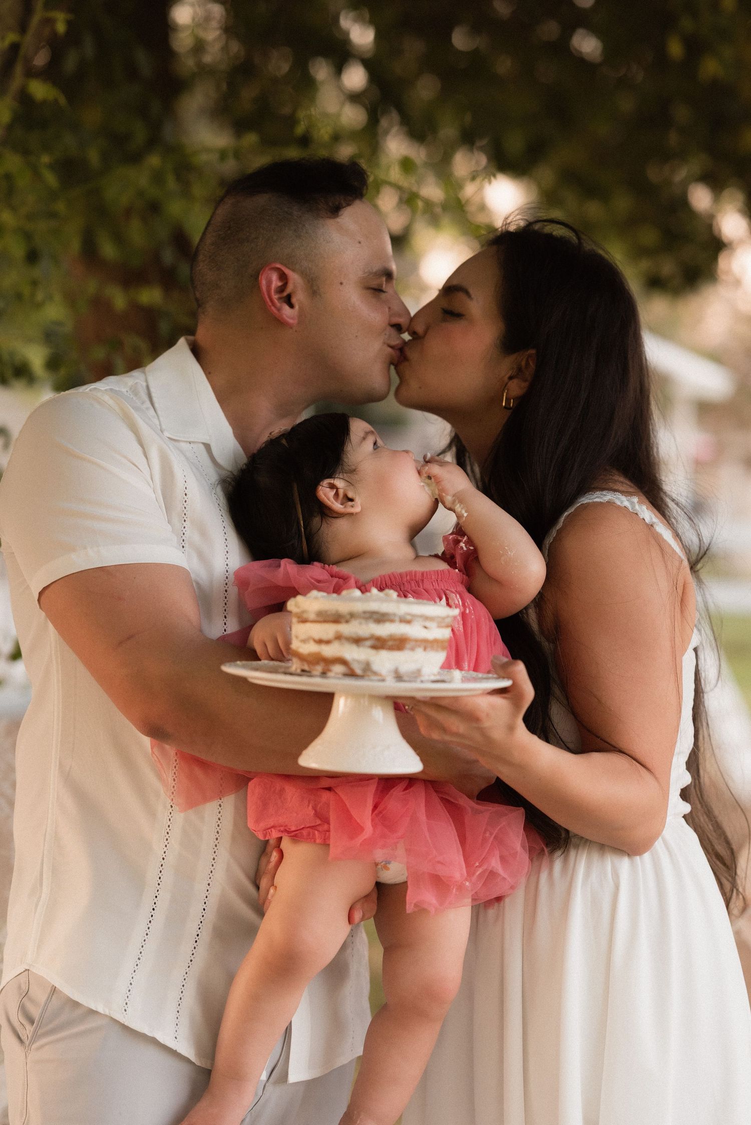 Parents kissing while holding baby girl in pink dress with smash cake at first birthday outdoor photo session.