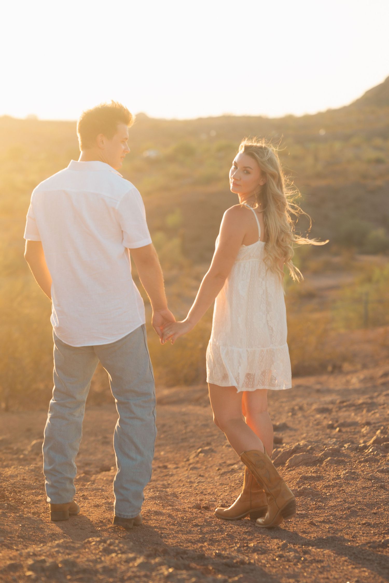 Couple holding hands at golden hour sunset in desert landscape, woman in white dress and boots looks back romantically.
