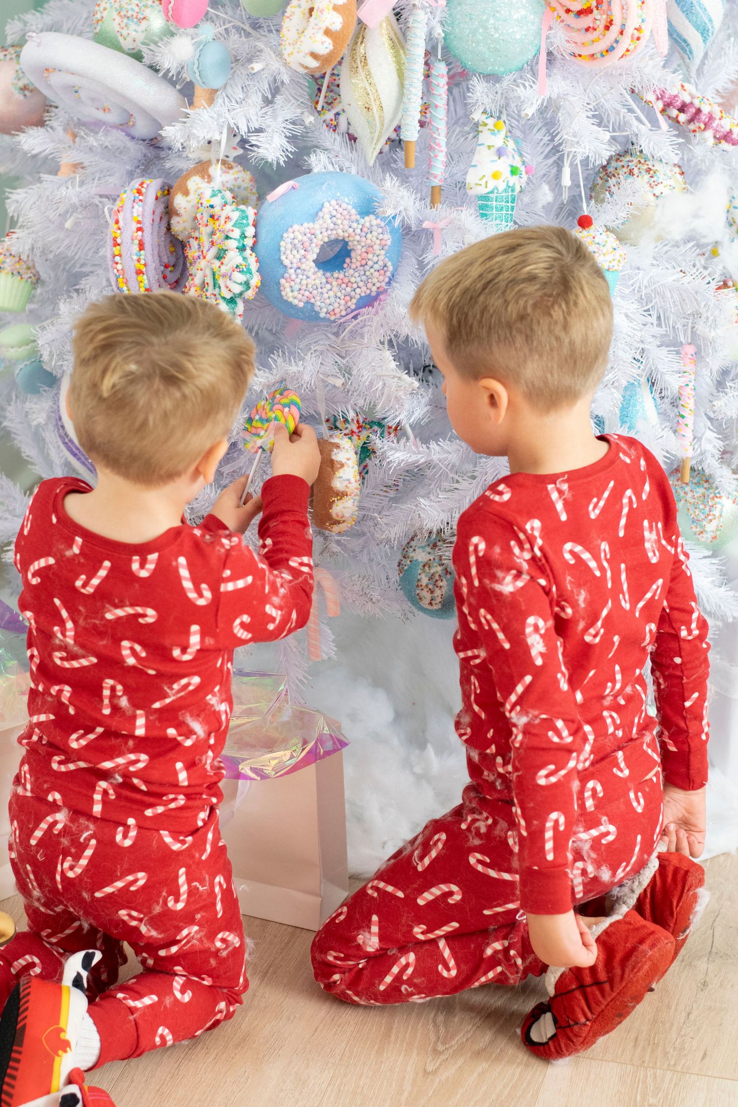 Two children in matching red candy cane pajamas decorating a white Christmas tree with colorful sweet-themed ornaments.