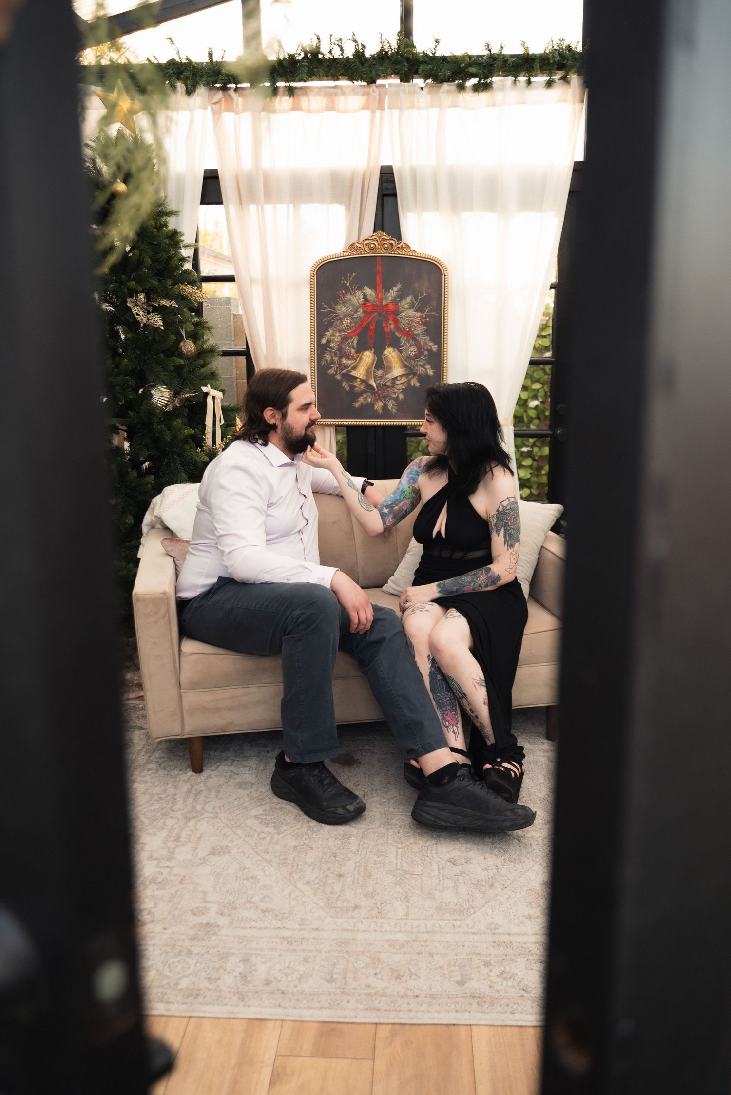 Two people sit together on a beige couch in a cozy sunlit room with sheer curtains and decorative wall art.