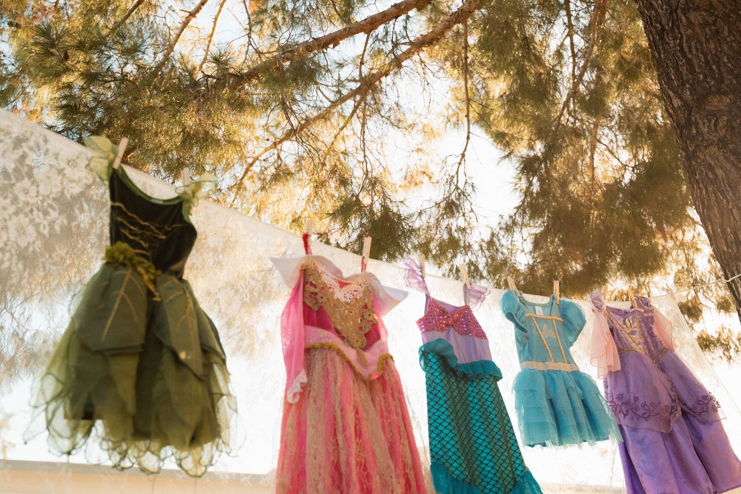Colorful girls' dresses hanging on a clothesline outdoors under pine trees, including green, pink, teal, and purple styles.