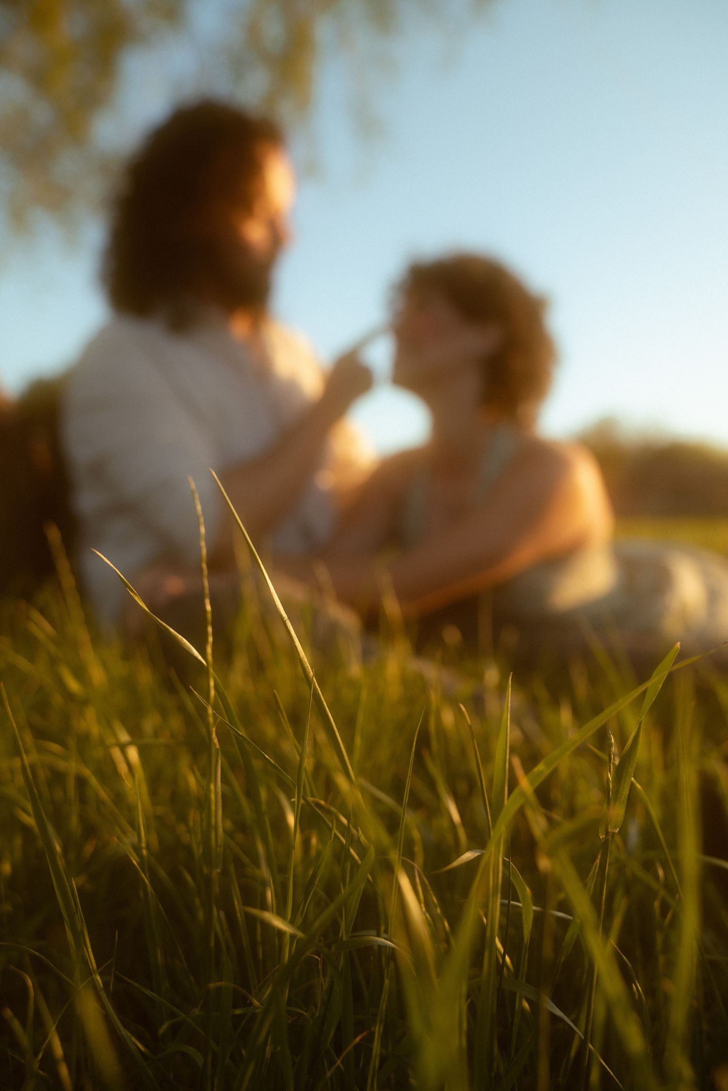 Couple sitting in tall green grass at golden hour sunset, sharing a romantic moment outdoors.