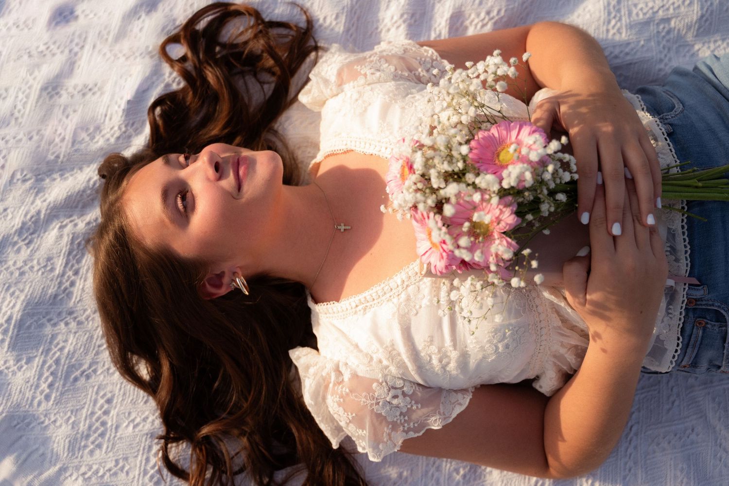 A young woman with long brown hair lies on white sand holding a bouquet of pink and white flowers, wearing a lace top.