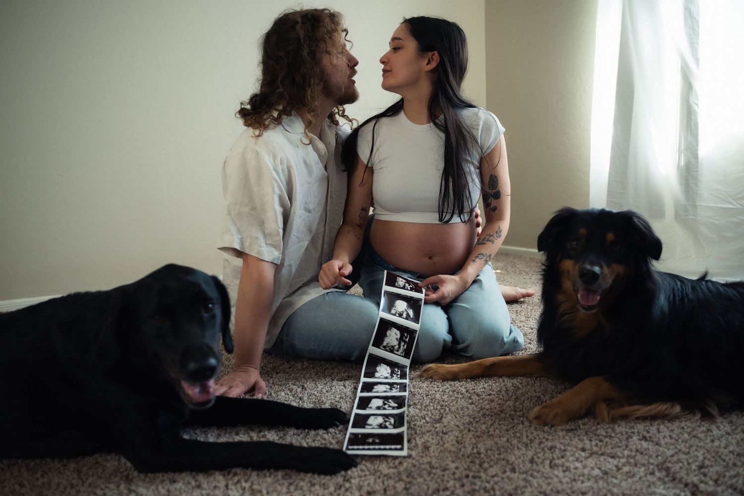 A couple and their black dog share an intimate moment on the floor while holding an ultrasound photo.