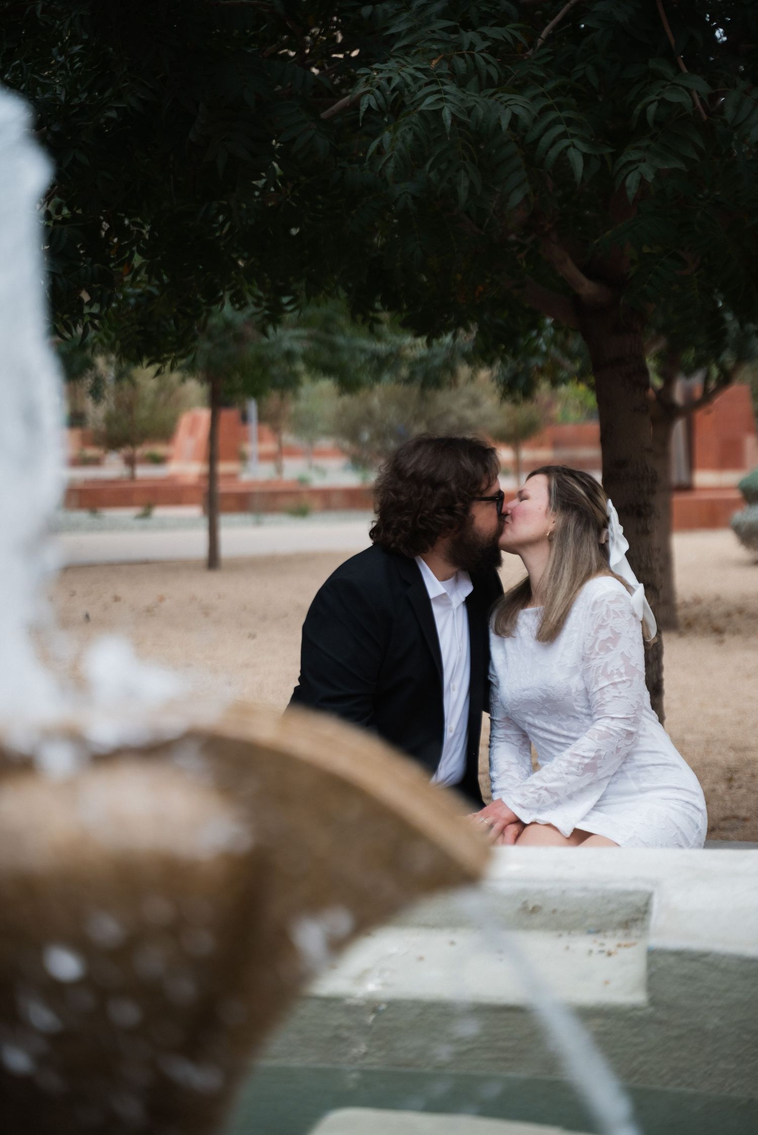A couple in white and black formal attire embracing on a bench surrounded by greenery.