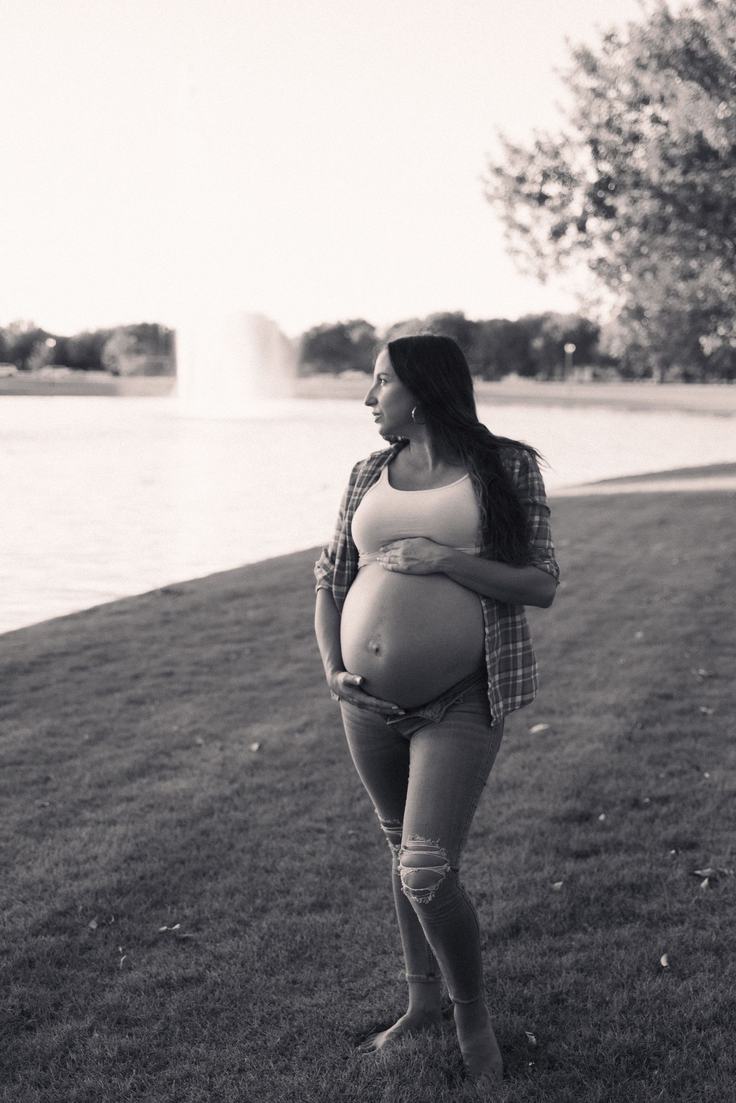 A black and white maternity photo shows an expectant mother in a plaid shirt and jeans by a lake with a fountain.
