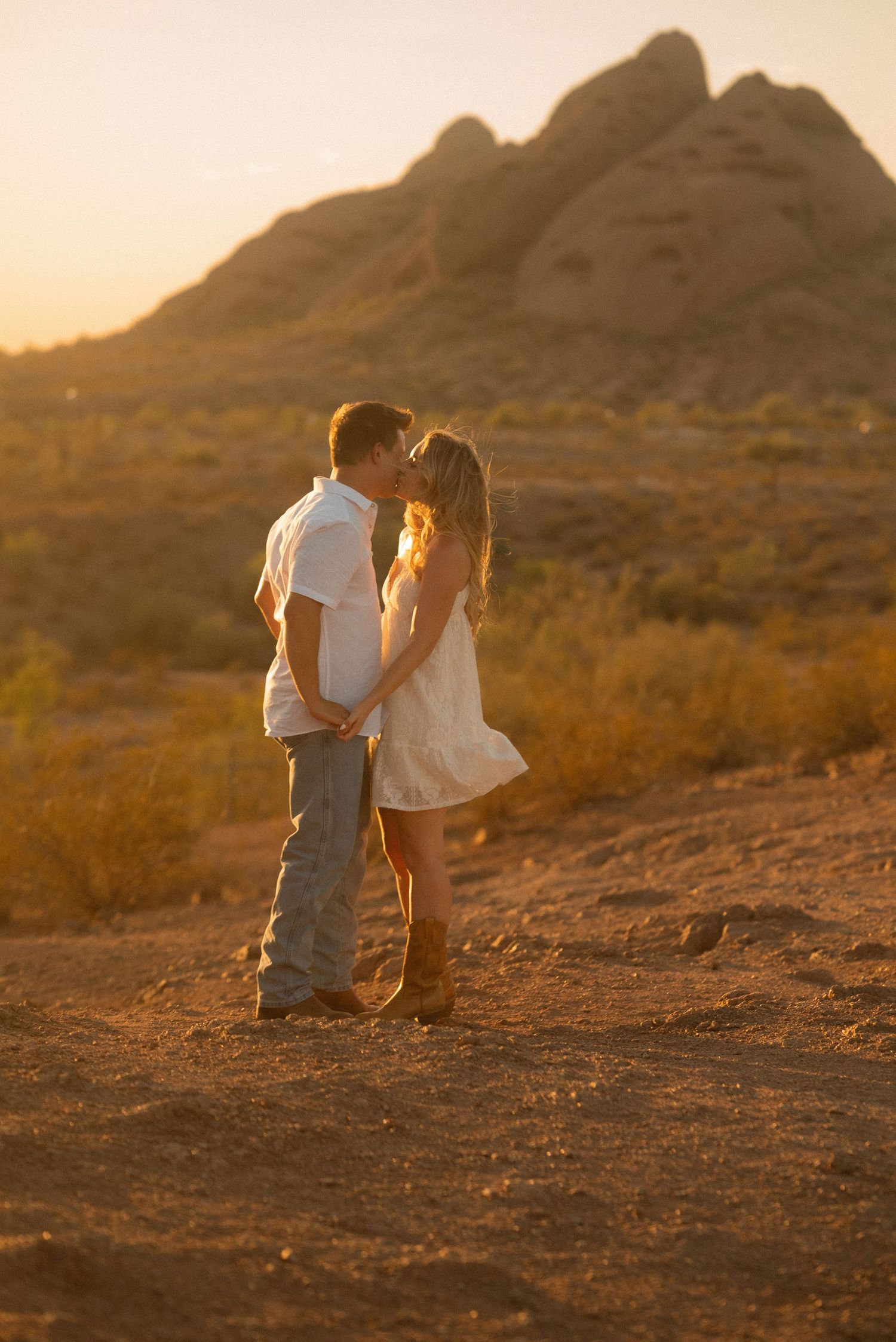 Couple sharing a romantic kiss at golden sunset in desert landscape with rocky mountain backdrop.