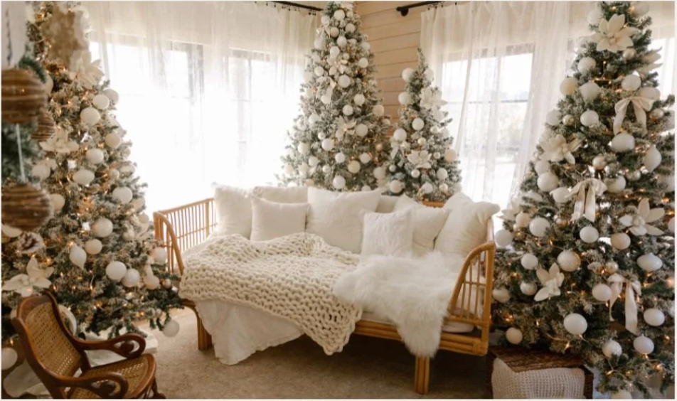 Elegant white Christmas living room with three flocked trees, gold ornaments, rattan daybed, and cozy knit throws.