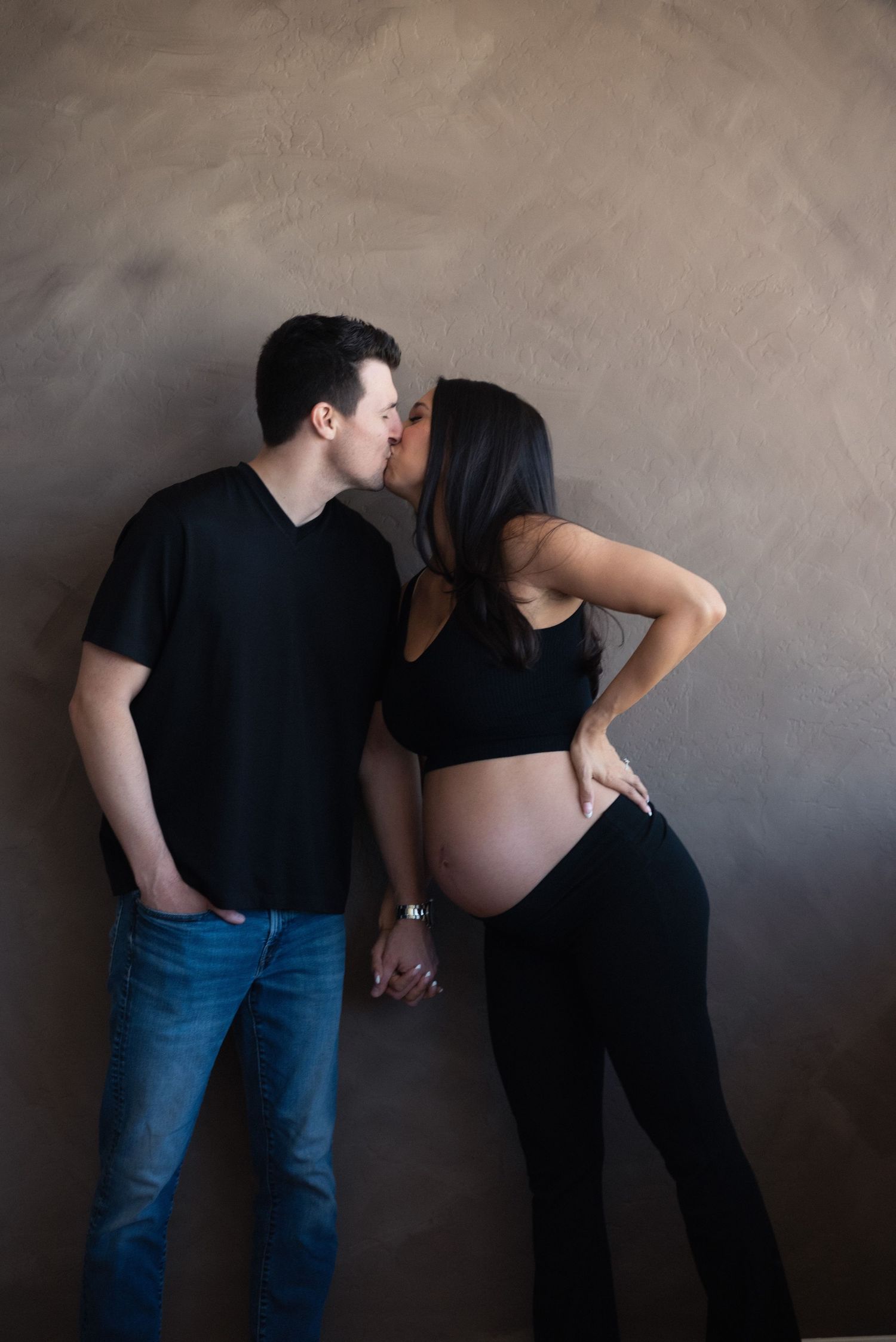 Couple sharing a kiss during a maternity photoshoot, woman in black crop top showing baby bump, man in black tee and jeans.