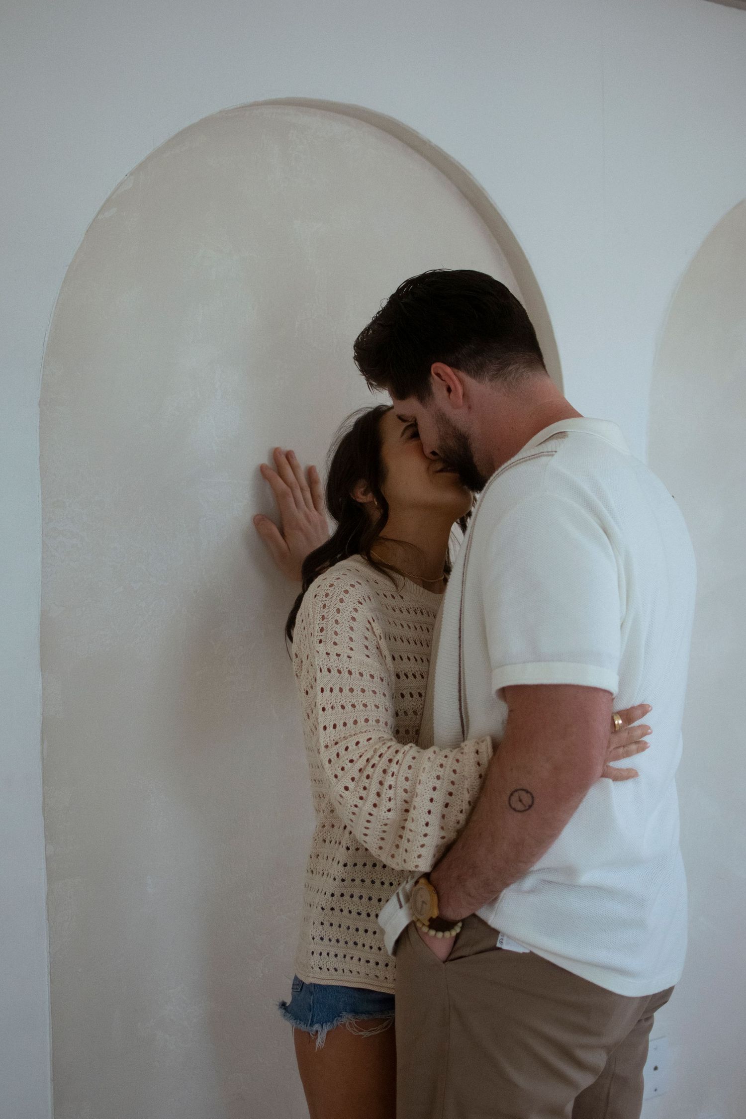 A couple embraces intimately against a white arched wall, with her wearing a knit sweater and him in a white t-shirt.