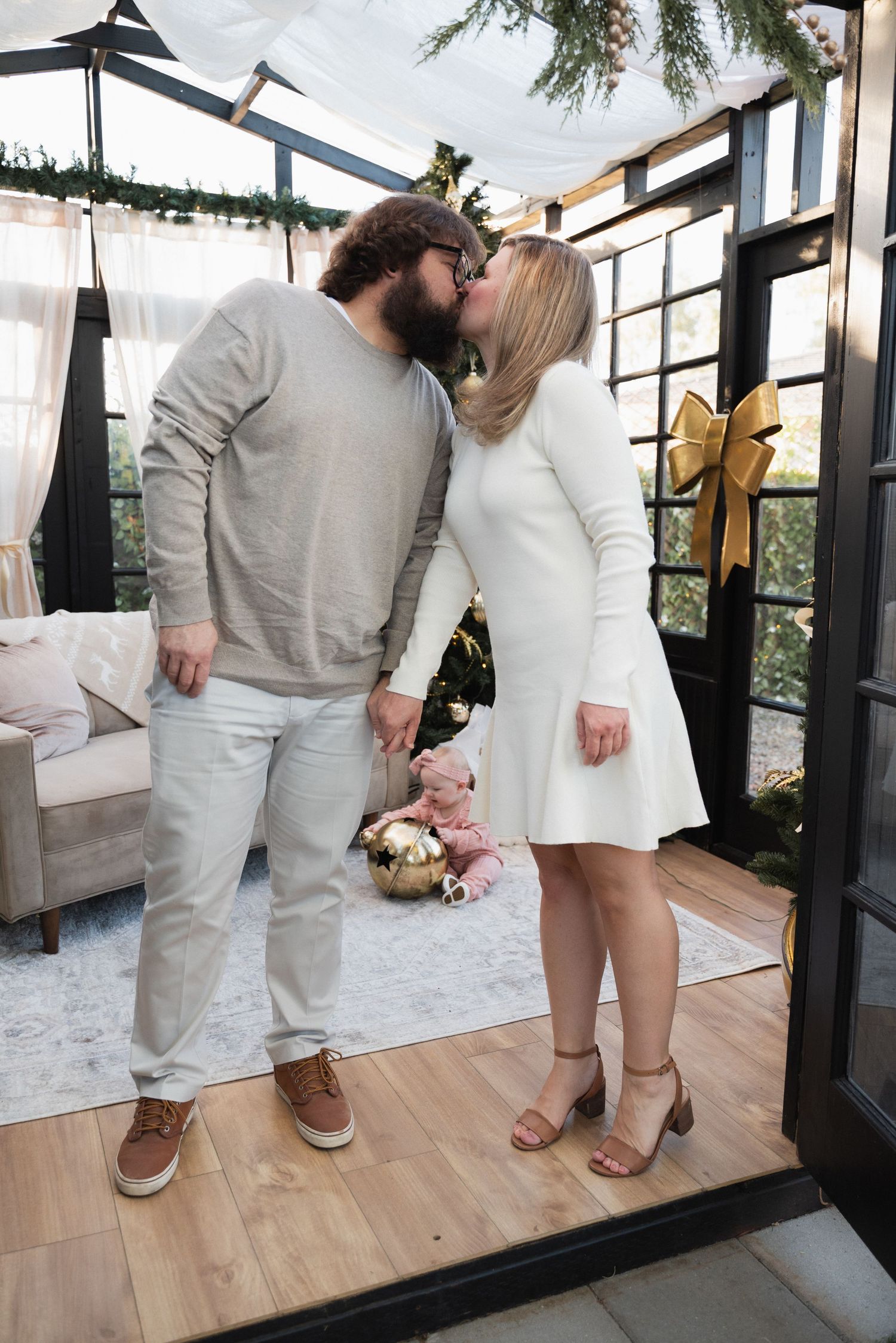 Couple kissing in decorated holiday greenhouse, man in gray sweater, woman in white dress, Christmas tree background.