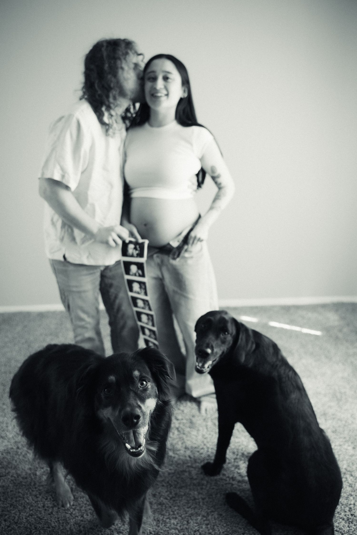 A couple embraces while their black dog sits beside them as they hold ultrasound images in a pregnancy announcement.