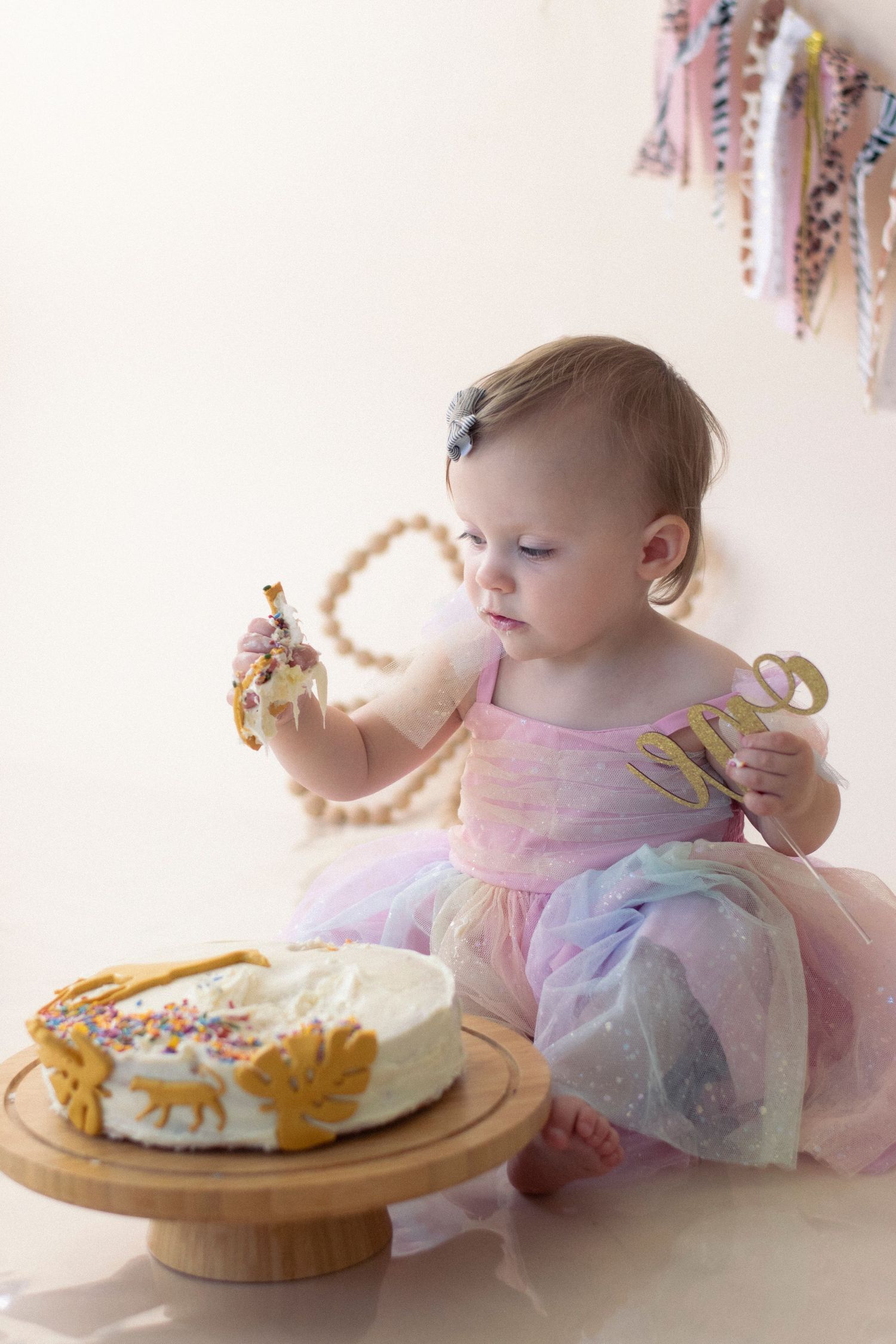 A toddler in a tulle dress sits near a birthday cake with sprinkles and decorations on a wooden board.
