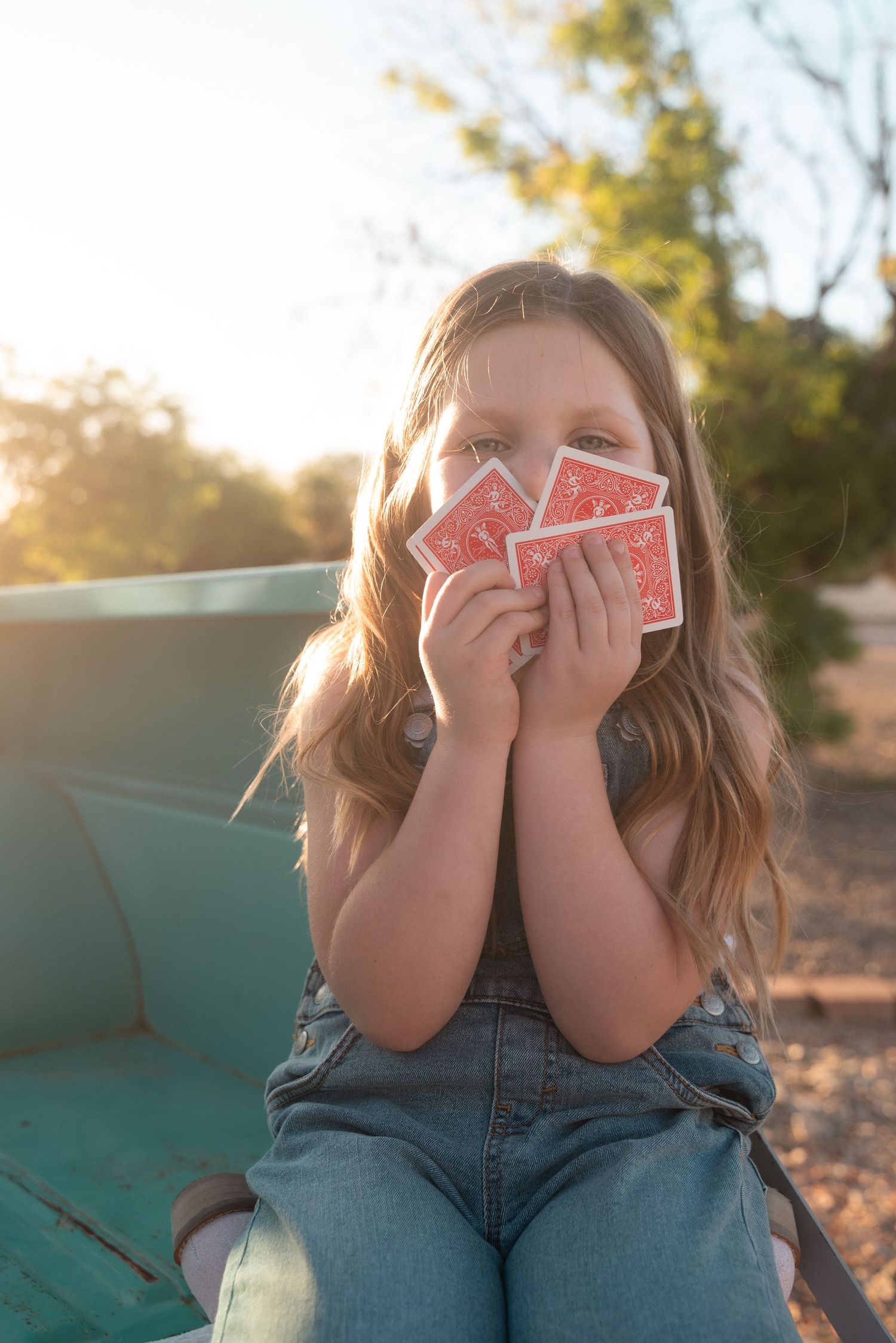 Young girl holding red playing cards in front of her face, sitting outdoors in golden sunlight.