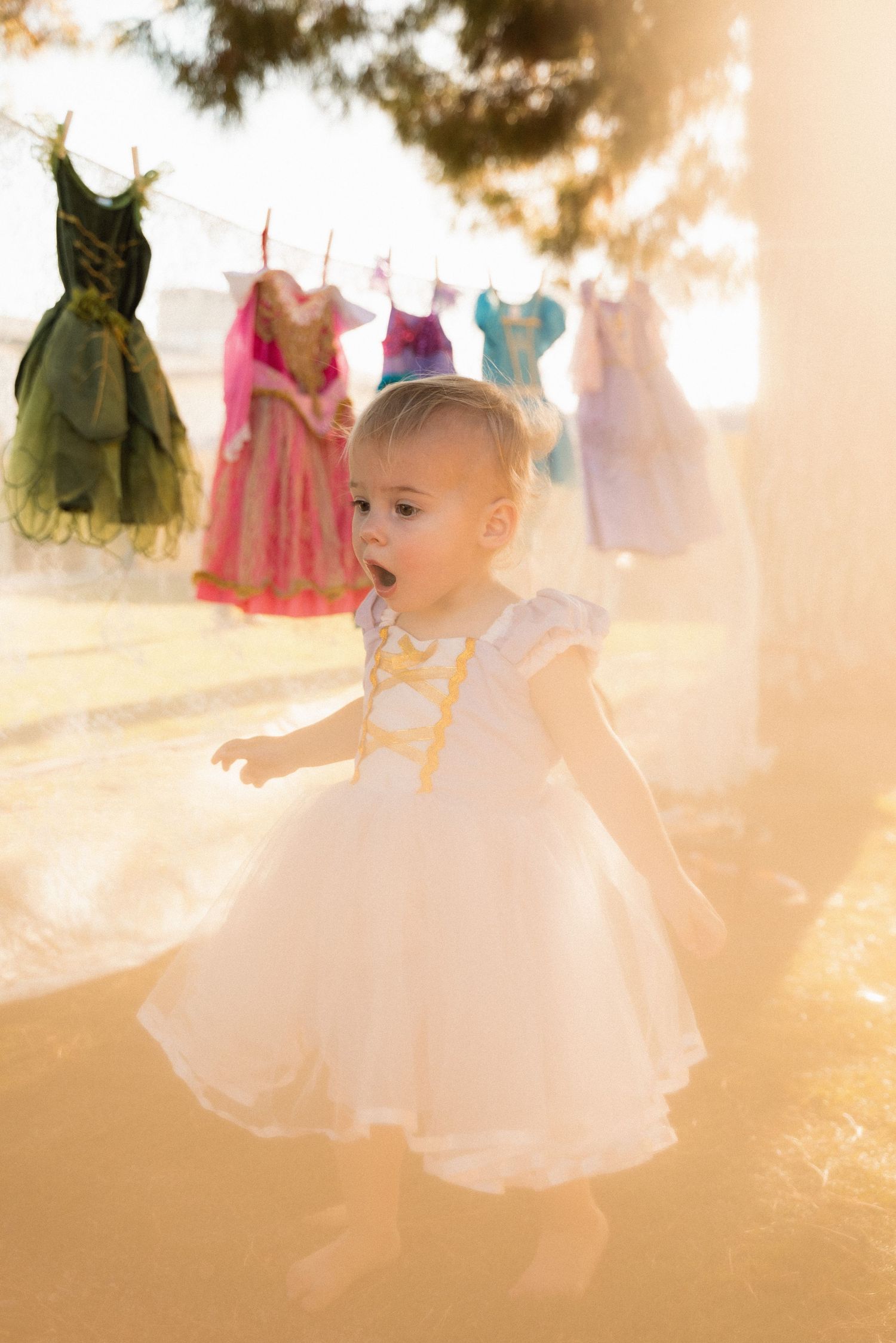 Toddler girl in white dress walks outdoors near colorful princess dresses hanging on a clothesline in golden sunlight.