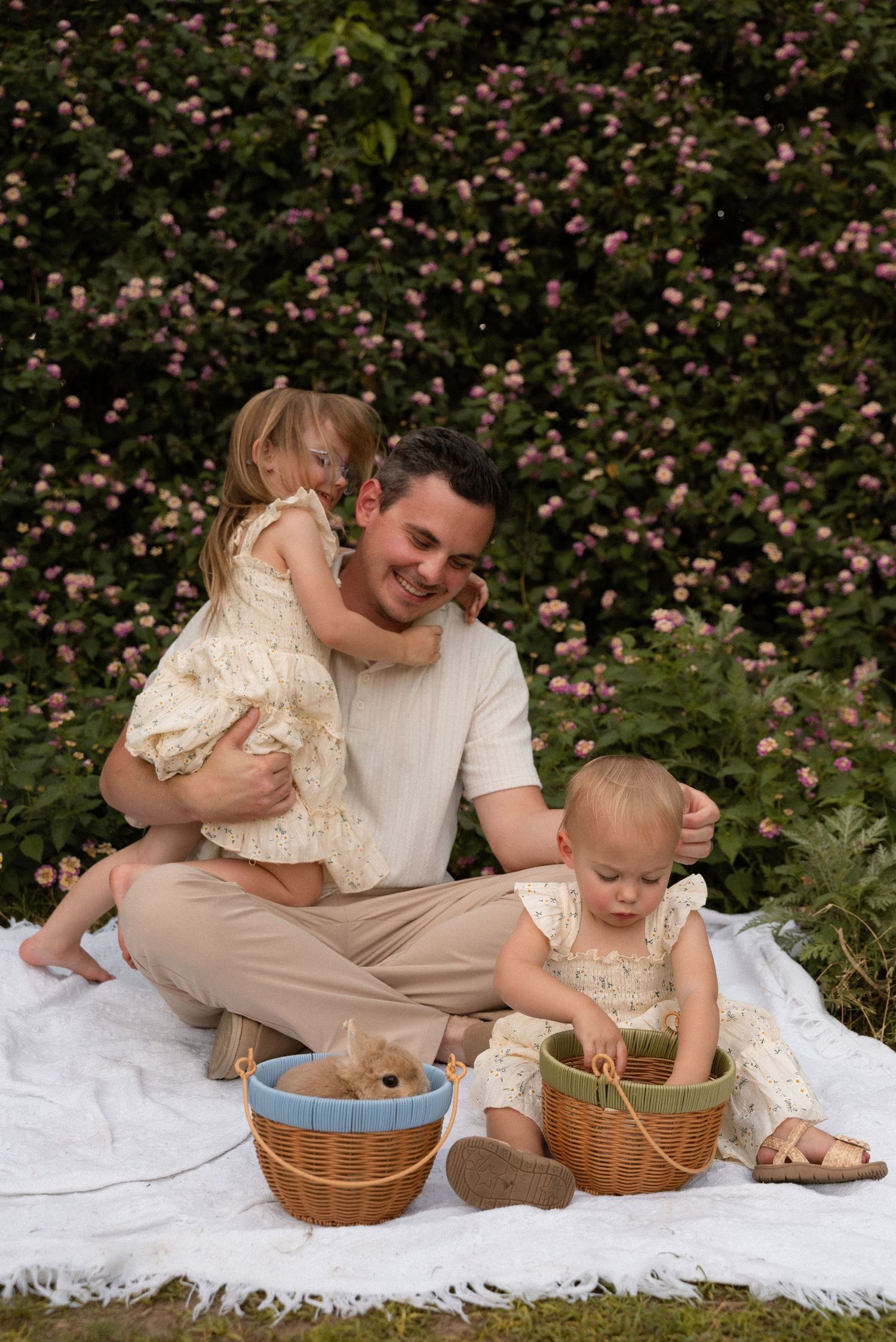 Father smiling with two toddler girls on a white blanket outdoors, surrounded by pink roses and wicker baskets.