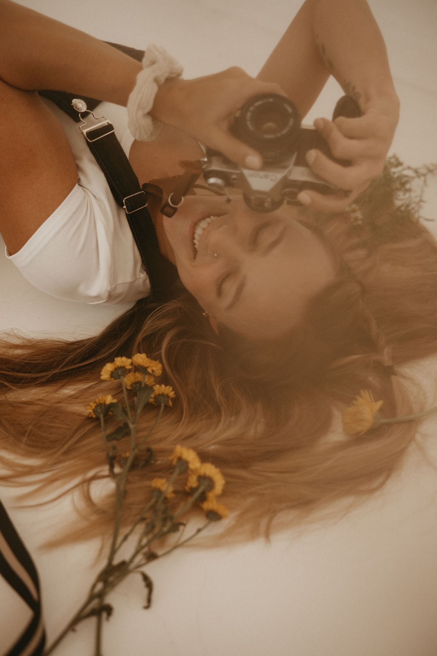 Smiling woman lying down holding a vintage camera surrounded by yellow wildflowers, wearing a white top with suspenders.