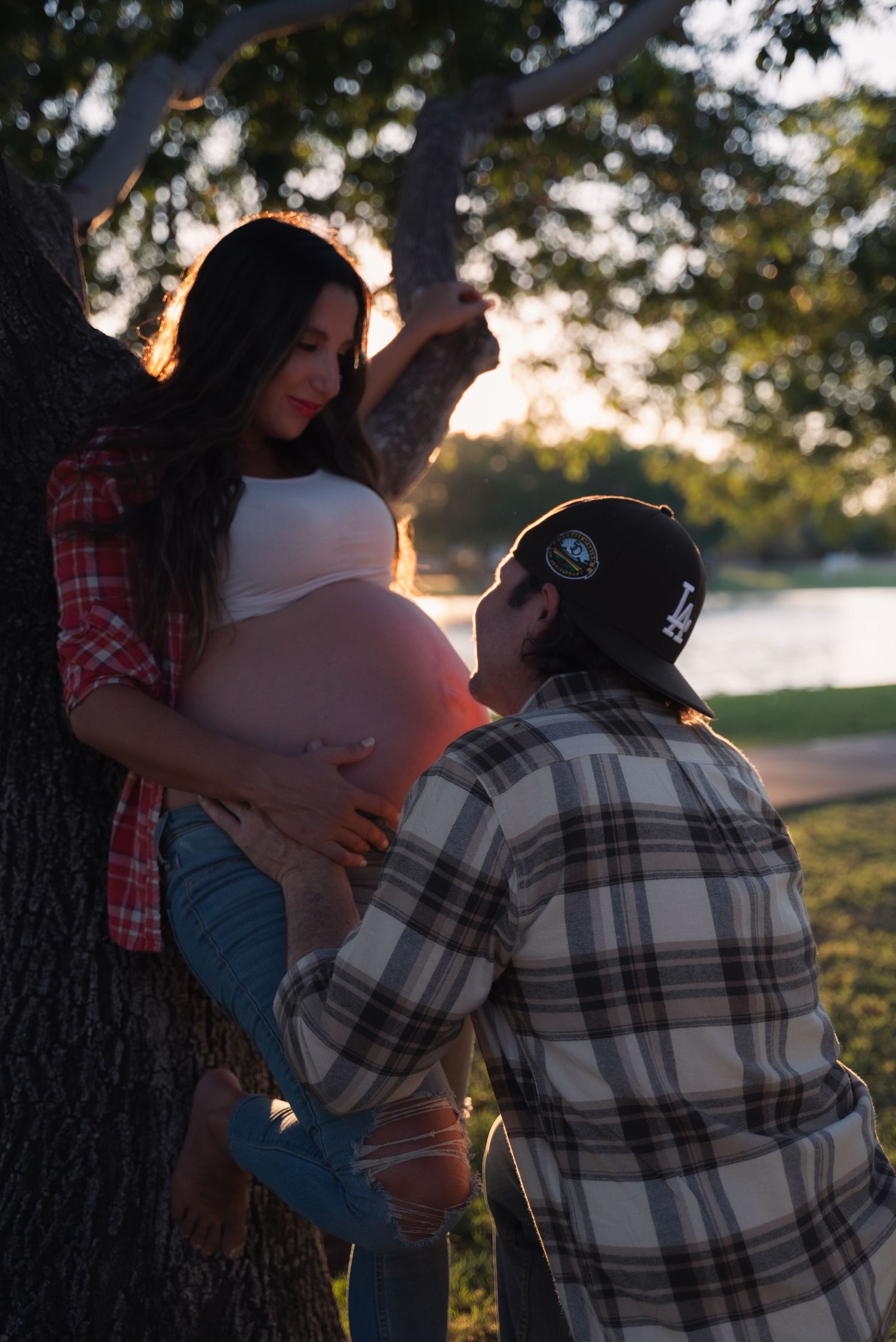 A couple shares an intimate maternity moment at sunset by a tree, with golden sunlight filtering through the branches.