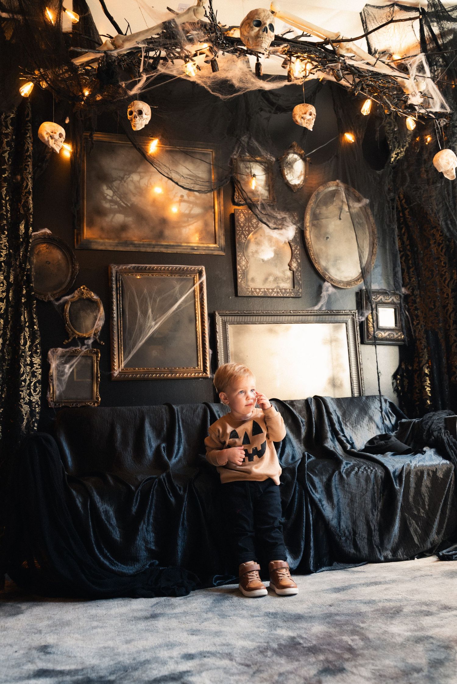 A spooky Halloween photo studio setup with black drapes, cobwebs, string lights and vintage frames on the wall.