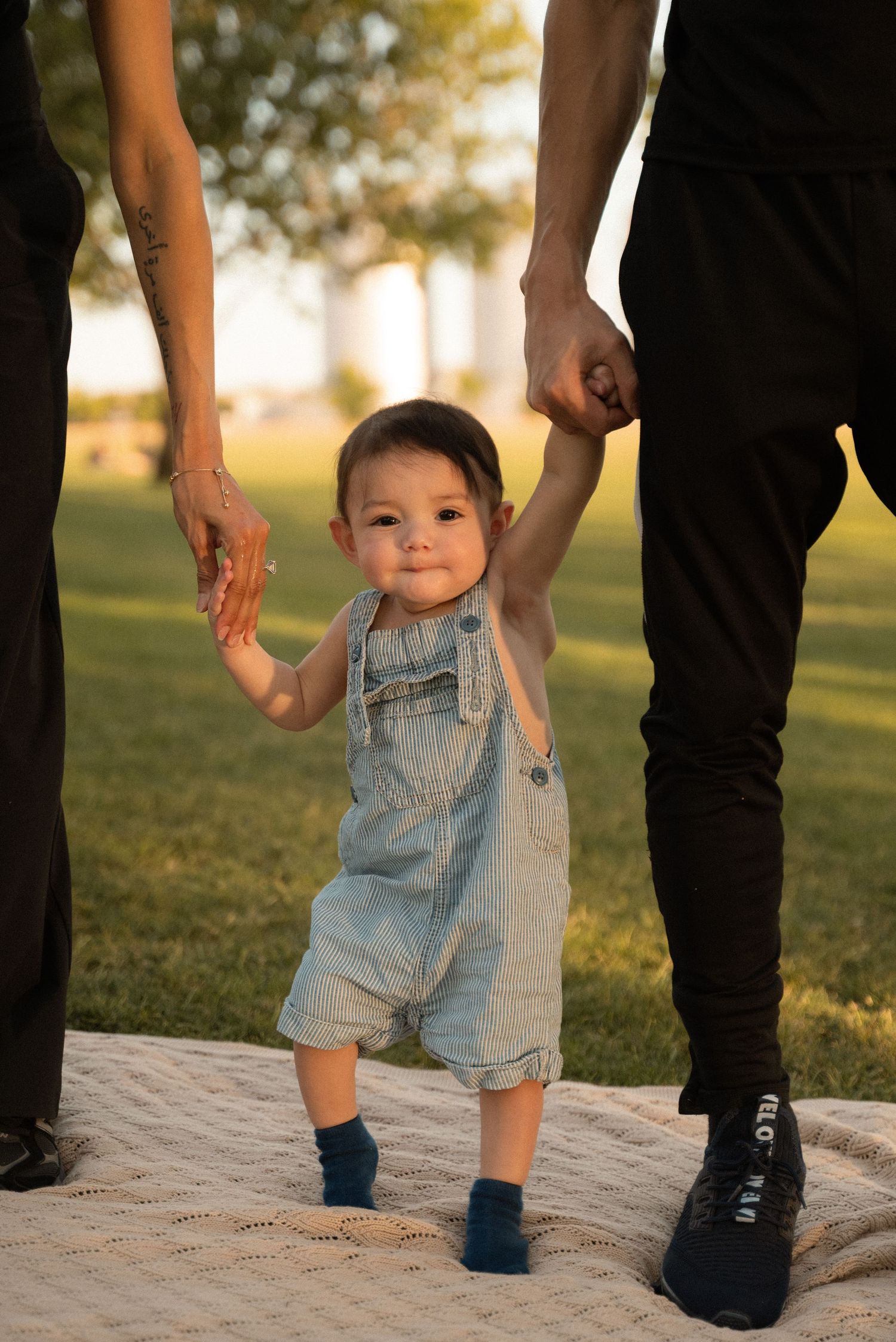 A baby in denim overalls takes first steps outdoors, held by parents hands on a sunny day.