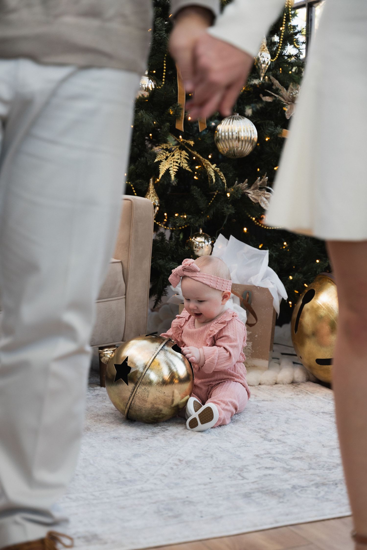 Baby in pink outfit sits by decorated Christmas tree with gold ornaments, parents standing nearby holding hands.