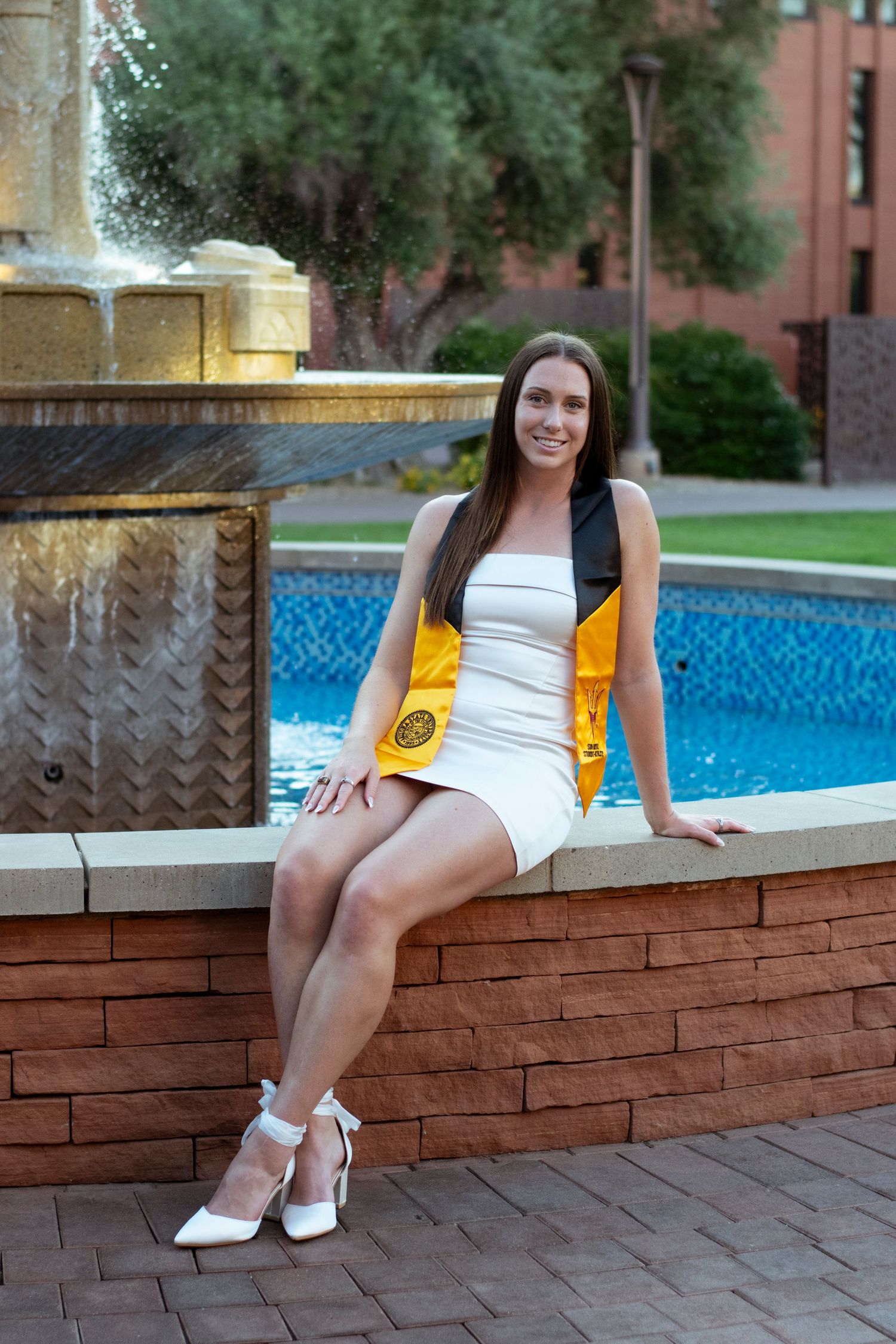 Person wearing a white dress and yellow sash sitting on a brick wall beside a swimming pool.