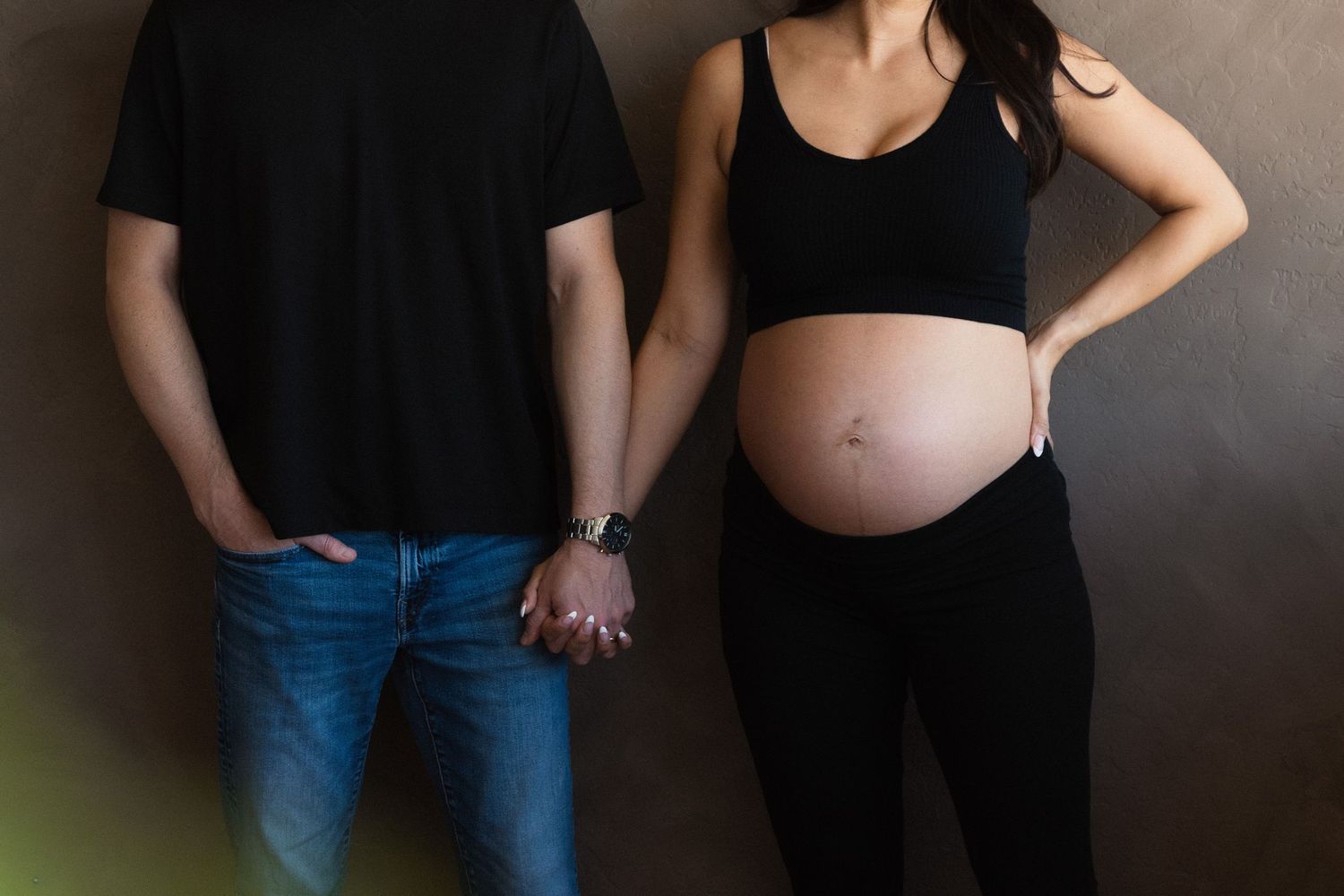 Pregnant woman in black outfit holds hands with partner against brown wall, showing baby bump.