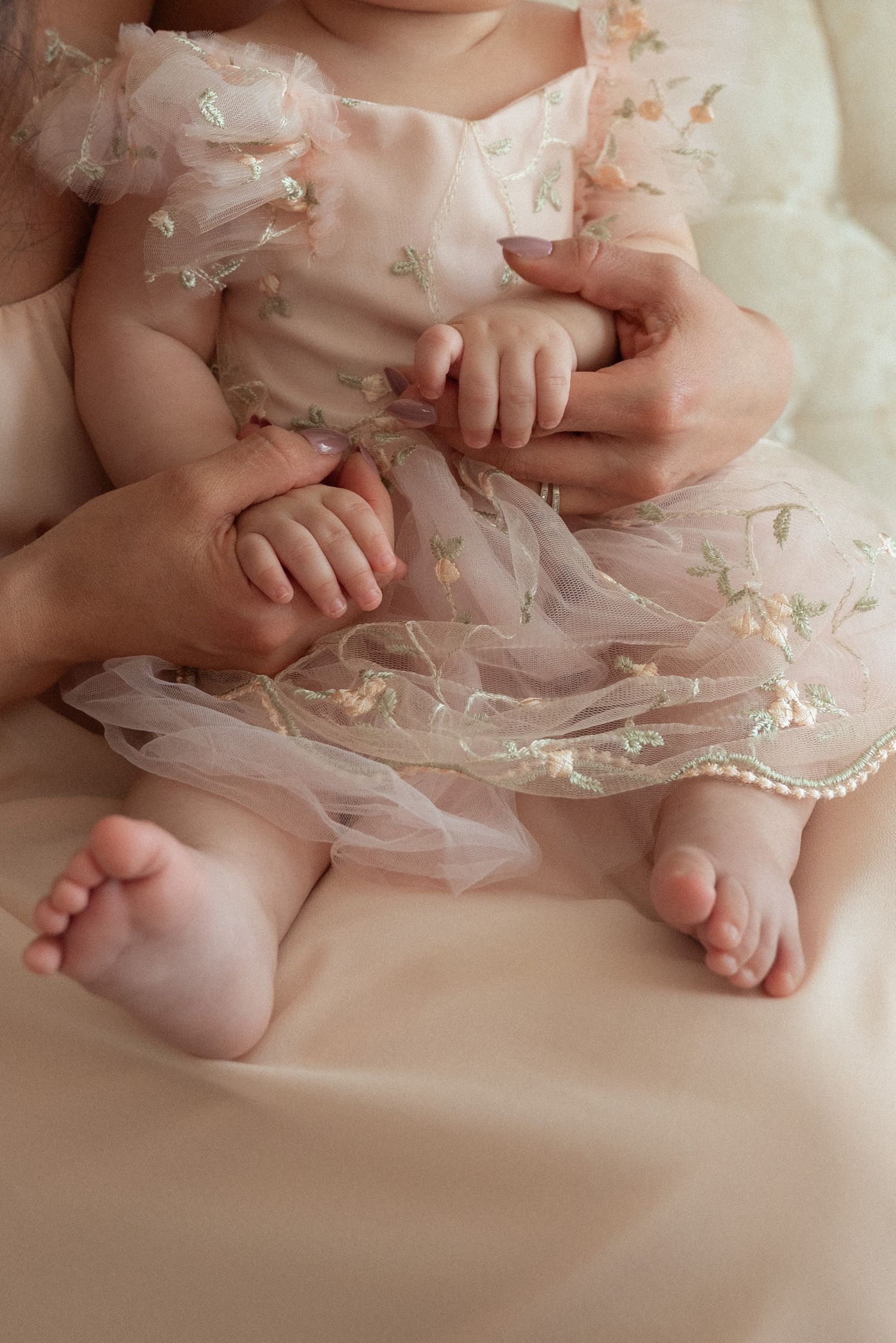 Baby in pink tulle dress held by mother, showing tiny hands and feet in a soft, tender moment.