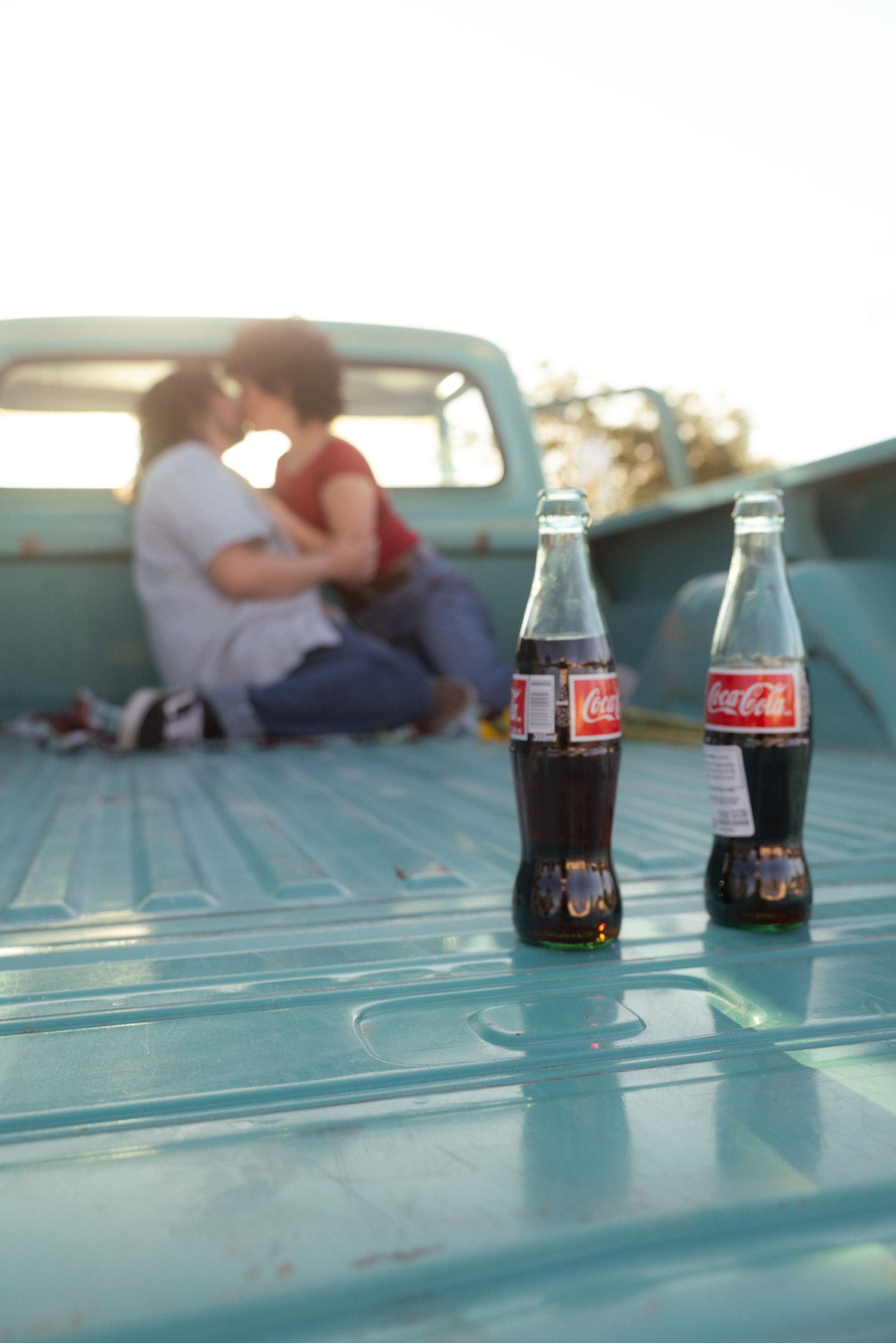 Two Coca-Cola glass bottles on a turquoise truck bed with a couple kissing in the background.