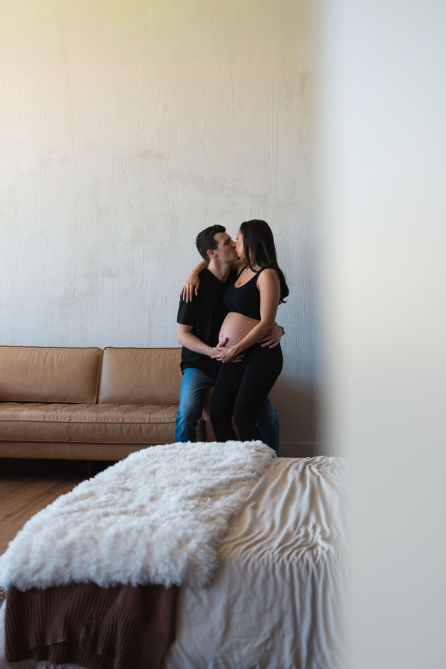 Couple sharing a tender kiss while cradling pregnant belly, standing in a cozy bedroom with leather sofa.