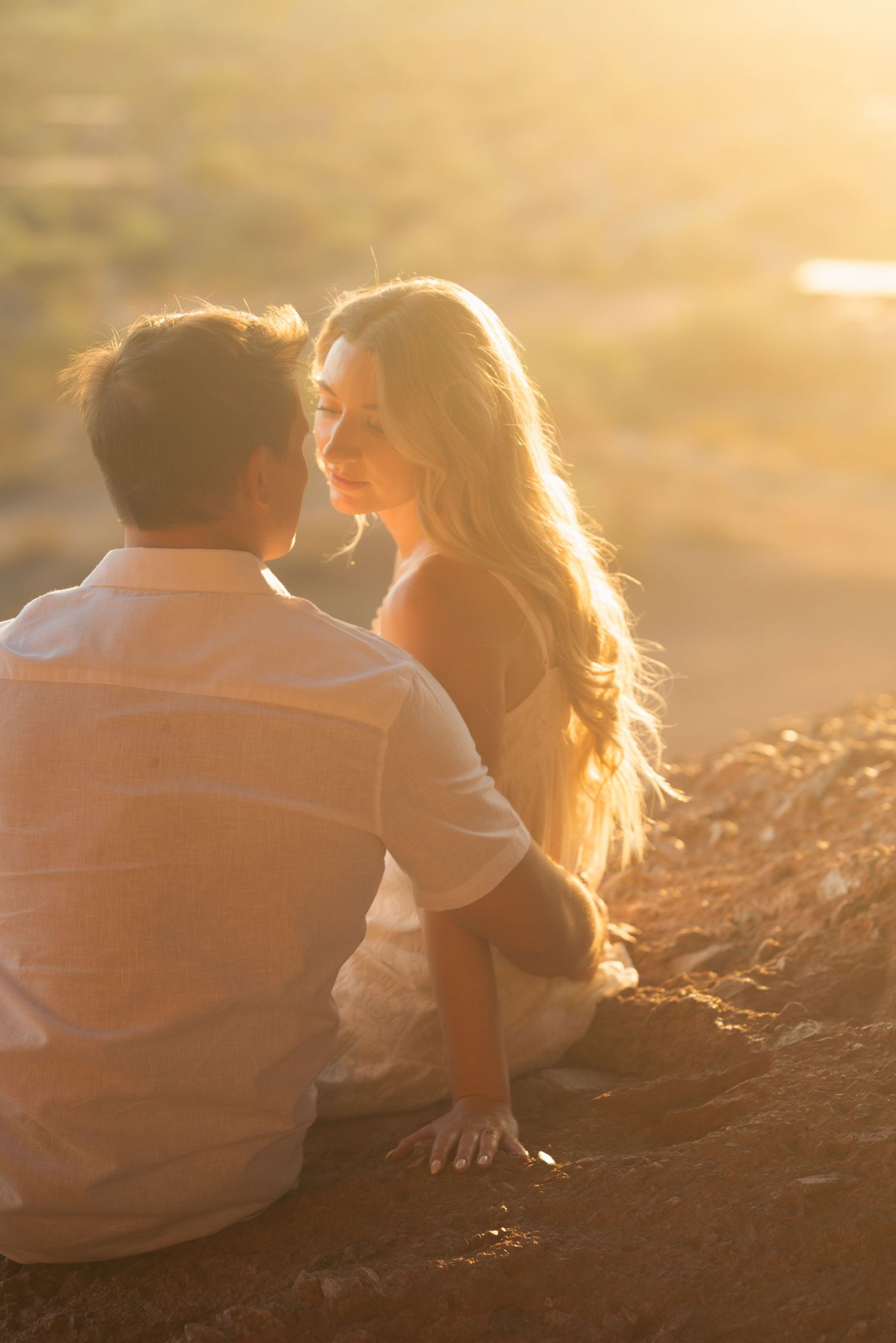 Couple sitting together on rocky ground at golden sunset, woman with long blonde hair gazing at man in white shirt.
