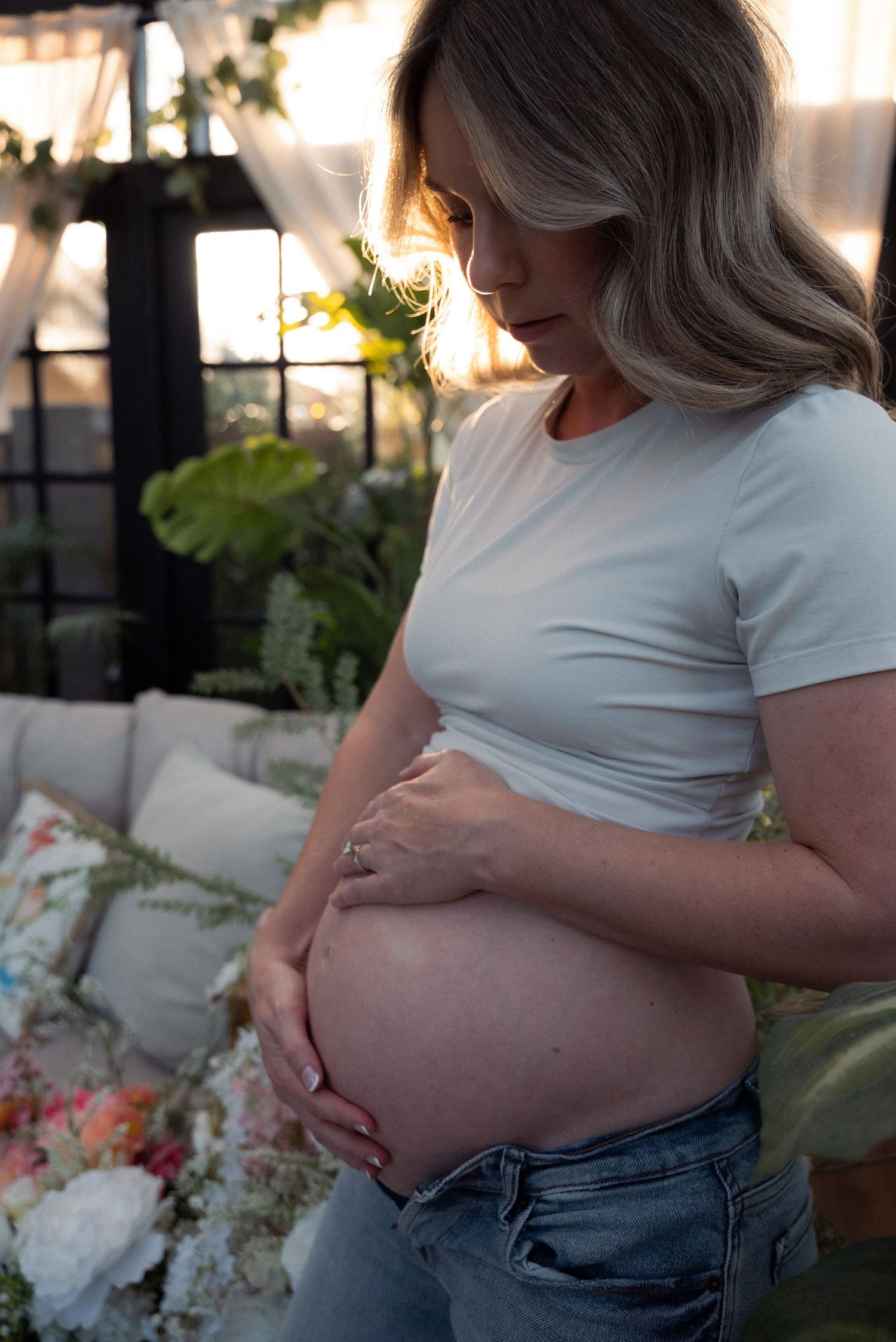 Side view of pregnant person in white shirt and jeans cradling belly against soft natural lighting.