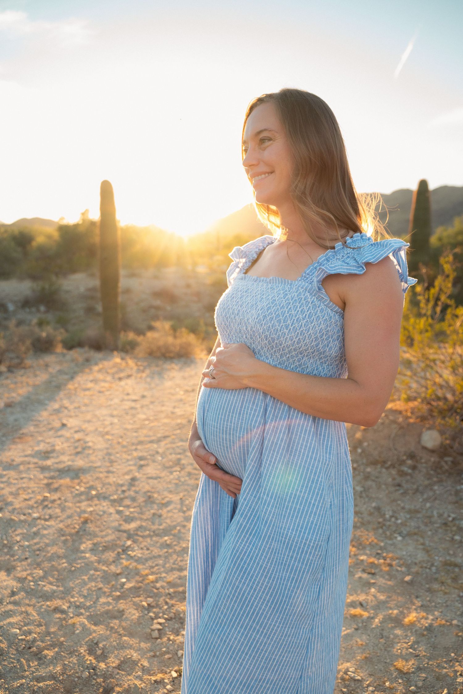 Expectant mother in blue dress poses in desert landscape at golden hour with saguaro cactus in background.