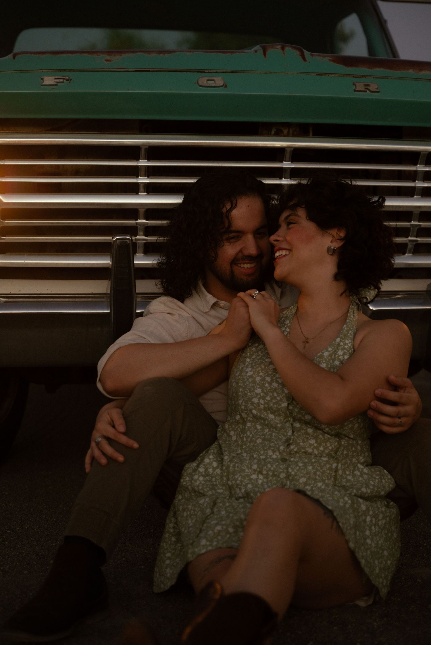 Couple sitting together laughing in front of a vintage teal Ford truck at dusk, woman in floral dress.