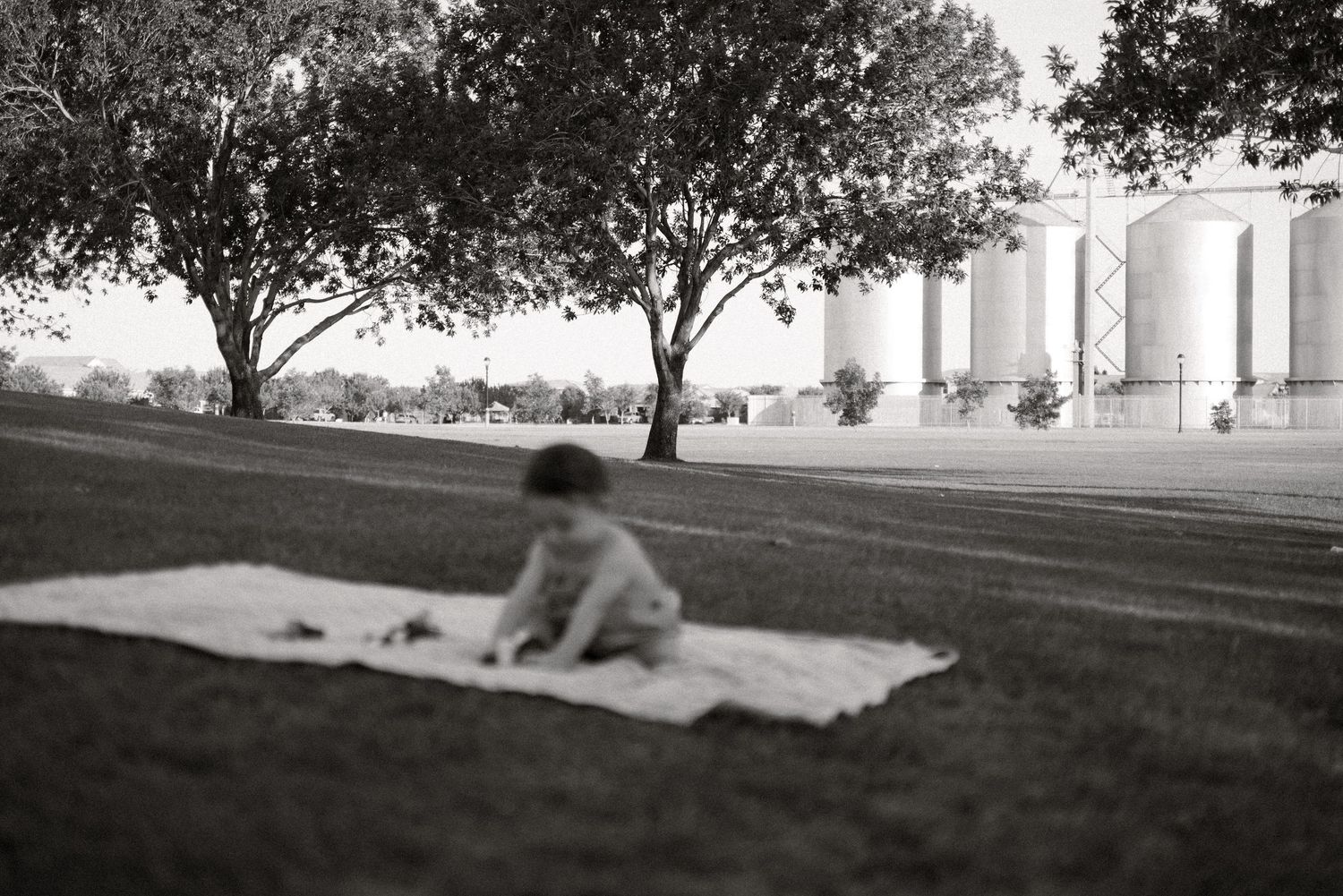 Black and white photo of a baby sitting on a blanket in a park with trees and grain silos in the background.