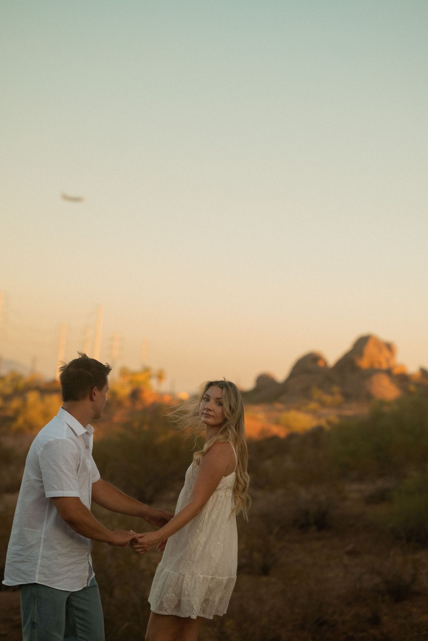 Couple holding hands at golden hour in desert landscape with rocky hills, woman in white dress looking back over shoulder.