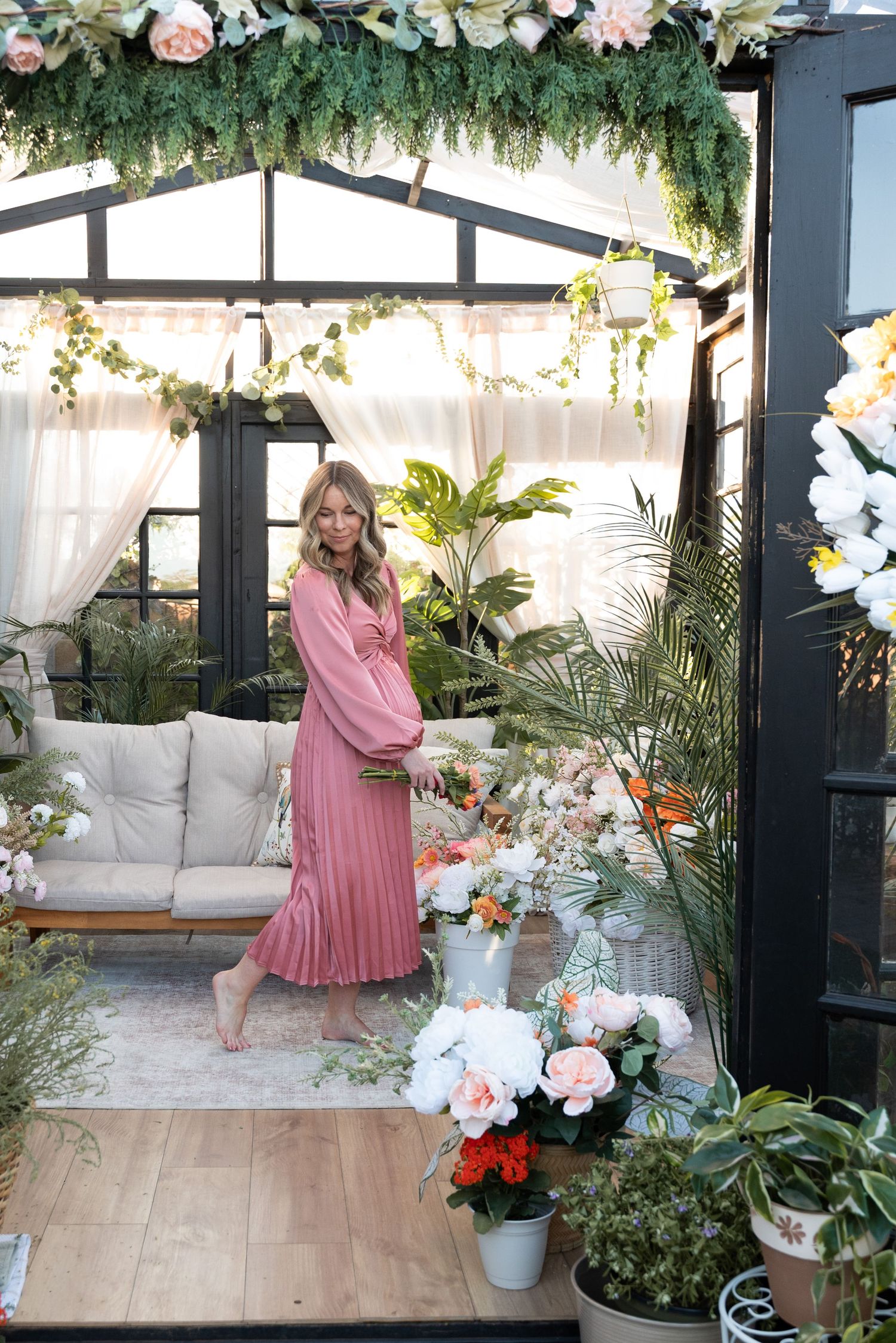 A greenhouse sunroom decorated with hanging greenery, floral arrangements, and white furniture creates a bright indoor garden oasis.
