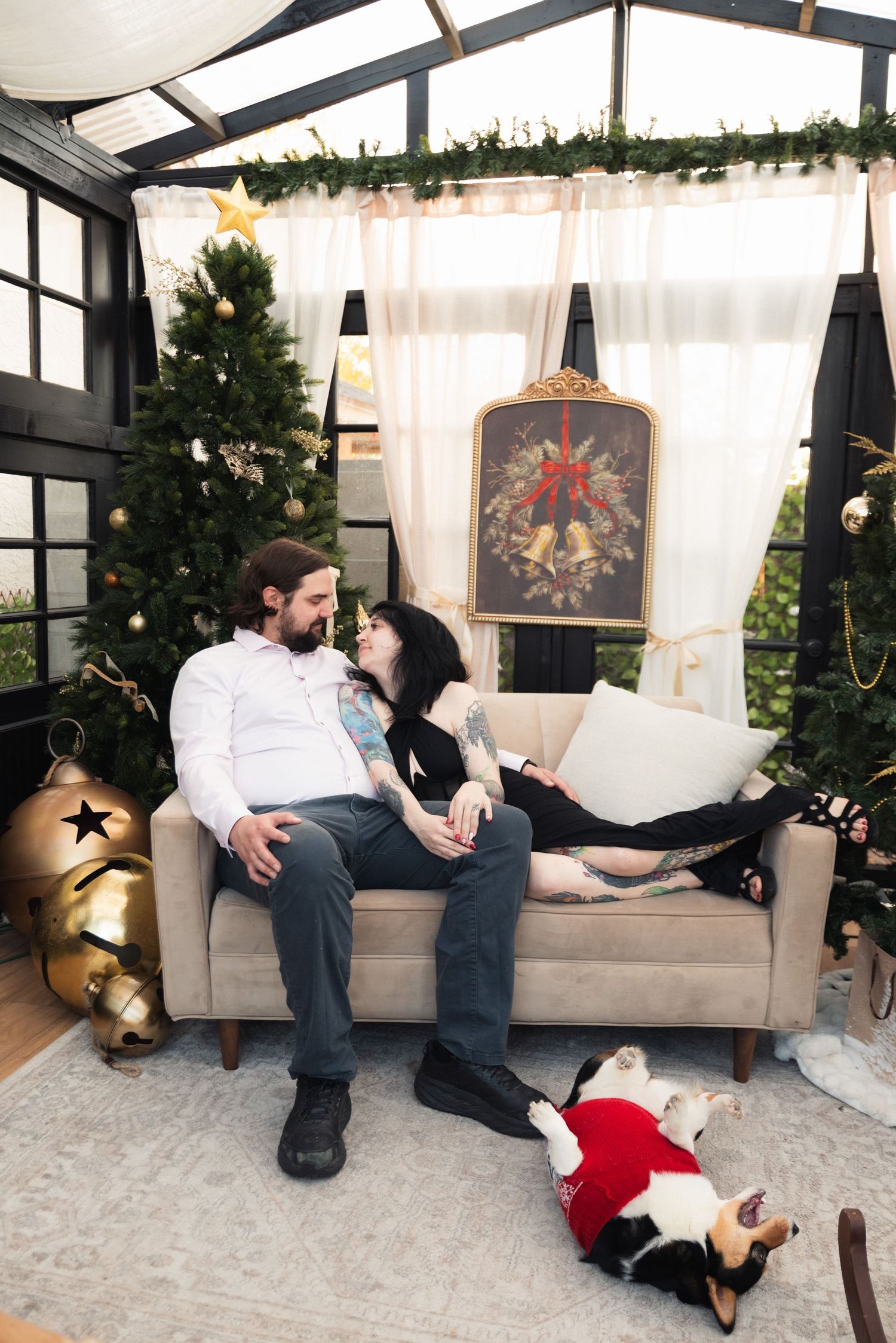 Couple relaxing on a beige sofa in a Christmas-decorated room with a tree, gold bells, and a dog in a red sweater.