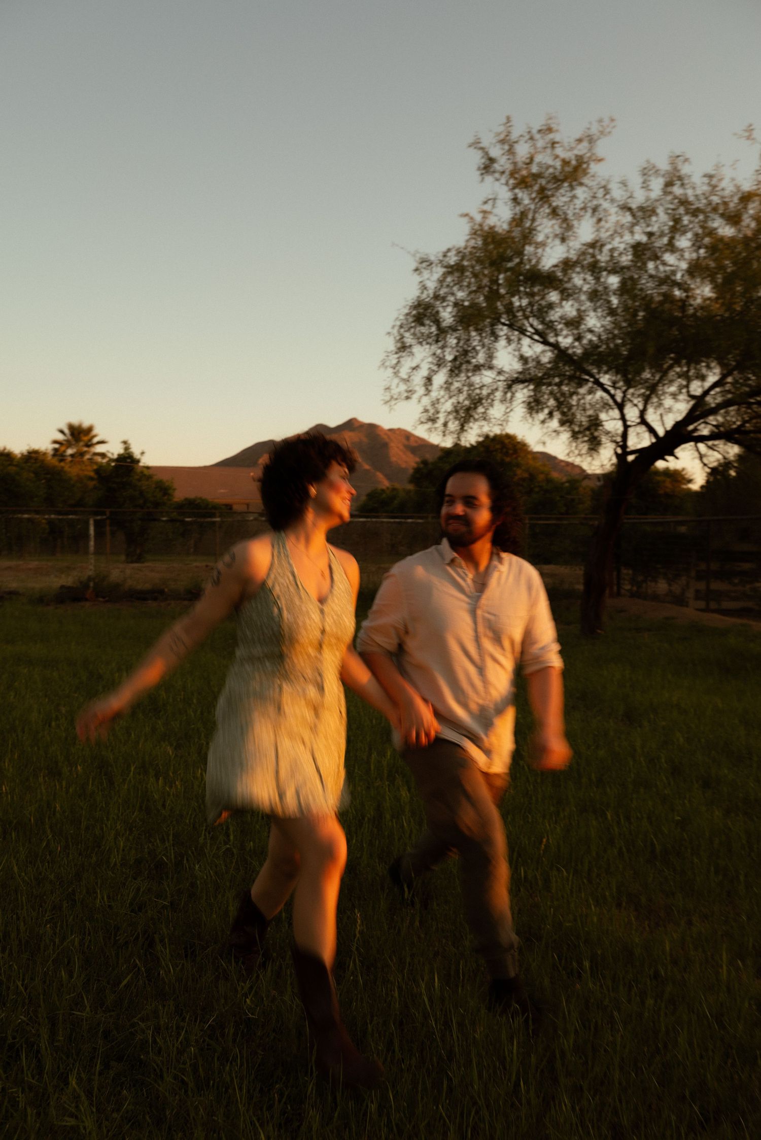 A couple dances joyfully in a green field at golden hour with mountains and a tree in the background.
