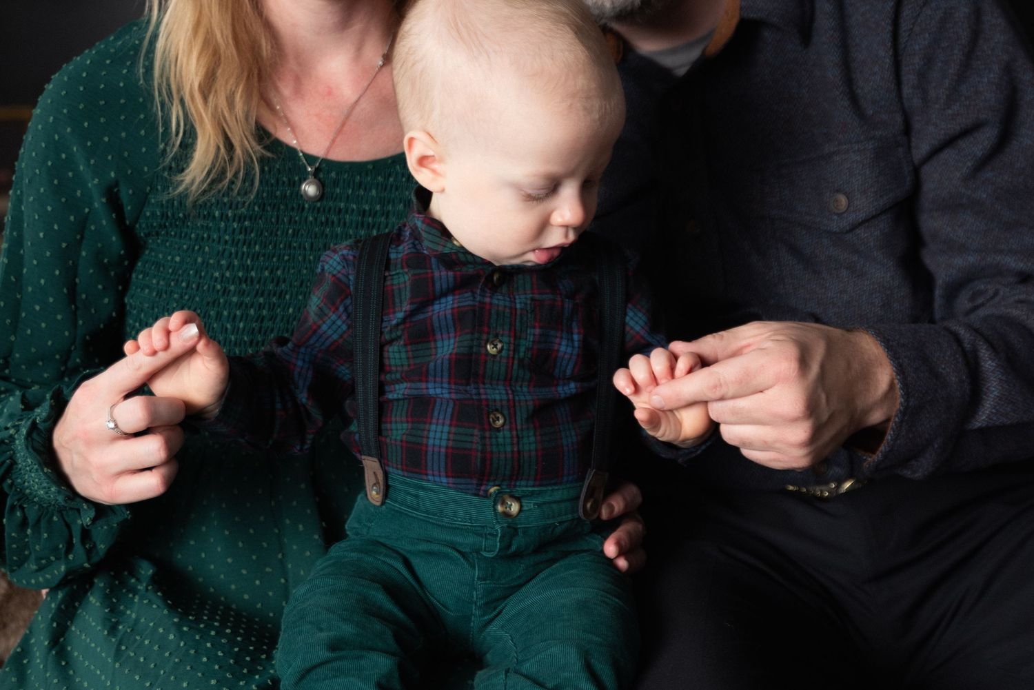 A family portrait of parents holding their baby boy dressed in plaid shirt and green pants in a studio setting.