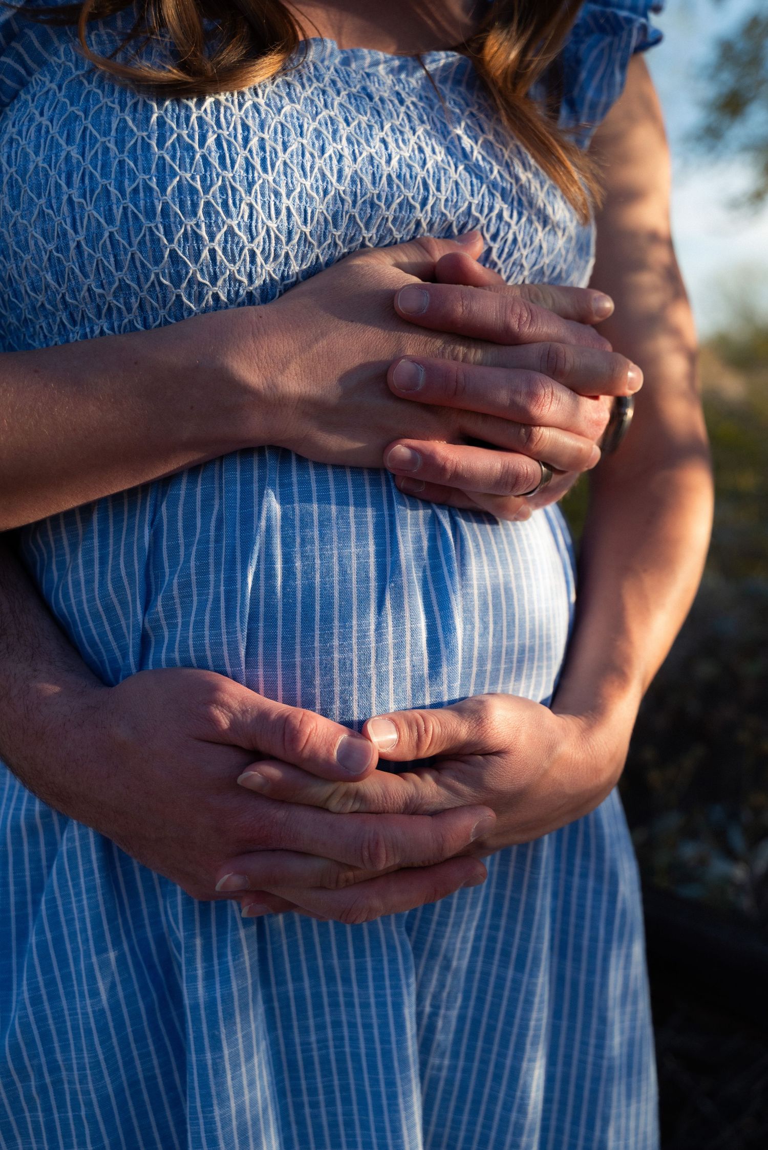 Close-up of hands cradling pregnant belly in blue striped dress with sunlit backdrop.