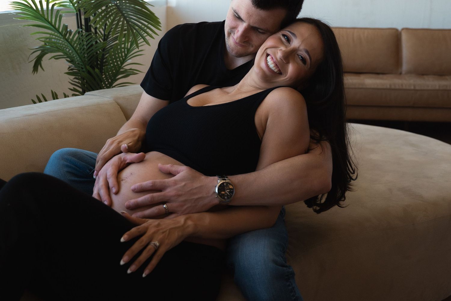 Pregnant woman in black dress smiles as partner embraces her from behind on a beige couch.