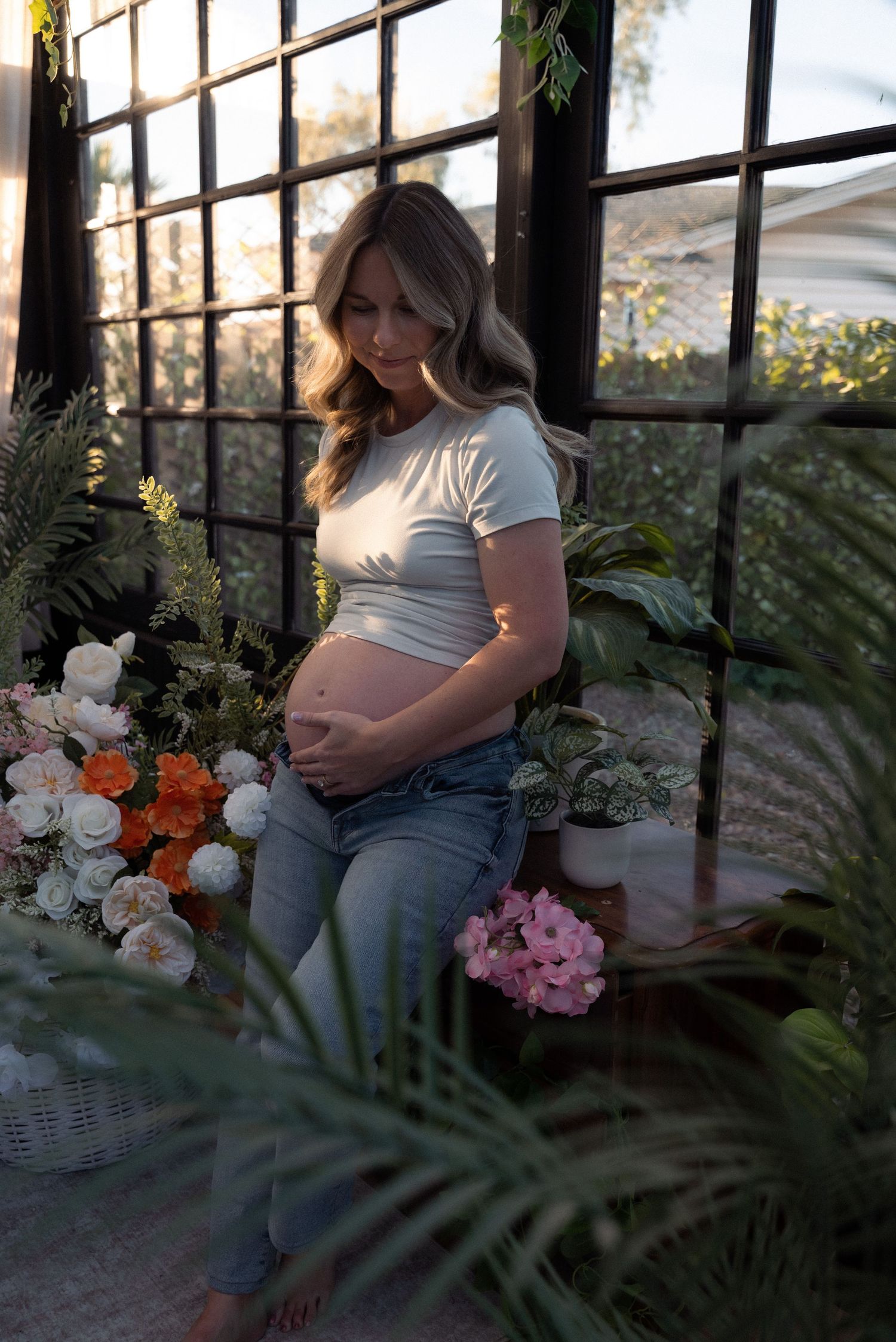 A serene maternity photoshoot in a sunlit greenhouse with lush plants and flowers creating a natural backdrop.