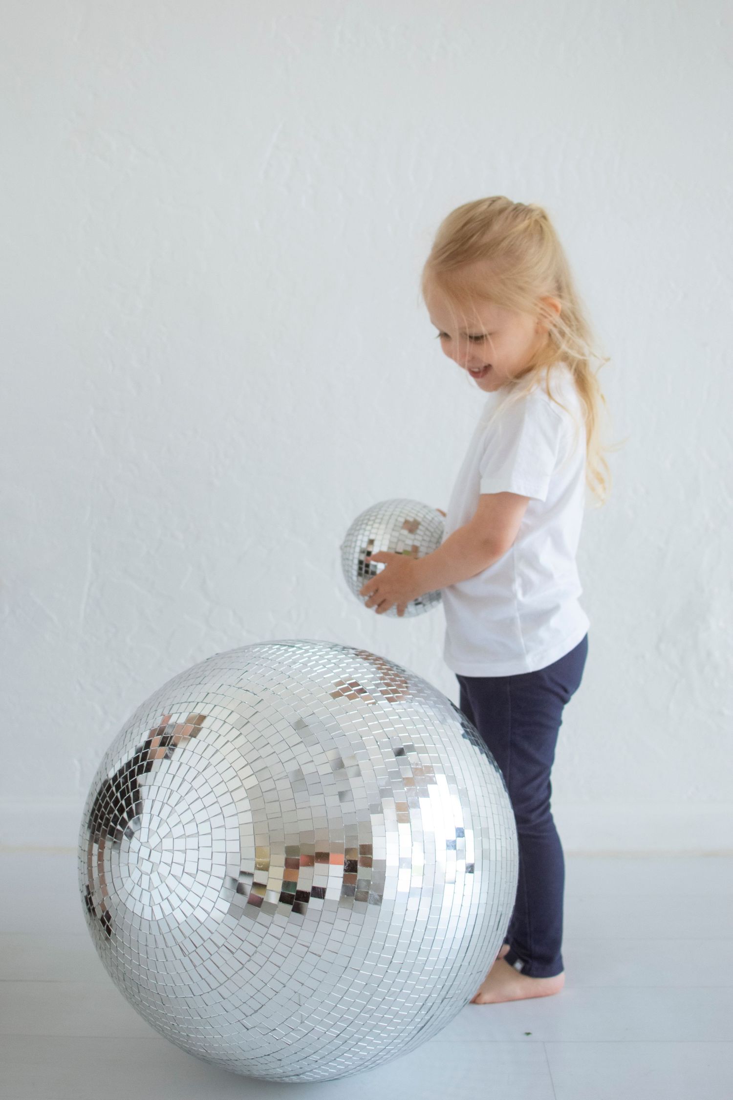 Small child in white shirt and navy shorts plays with large, shimmering disco ball on white floor.