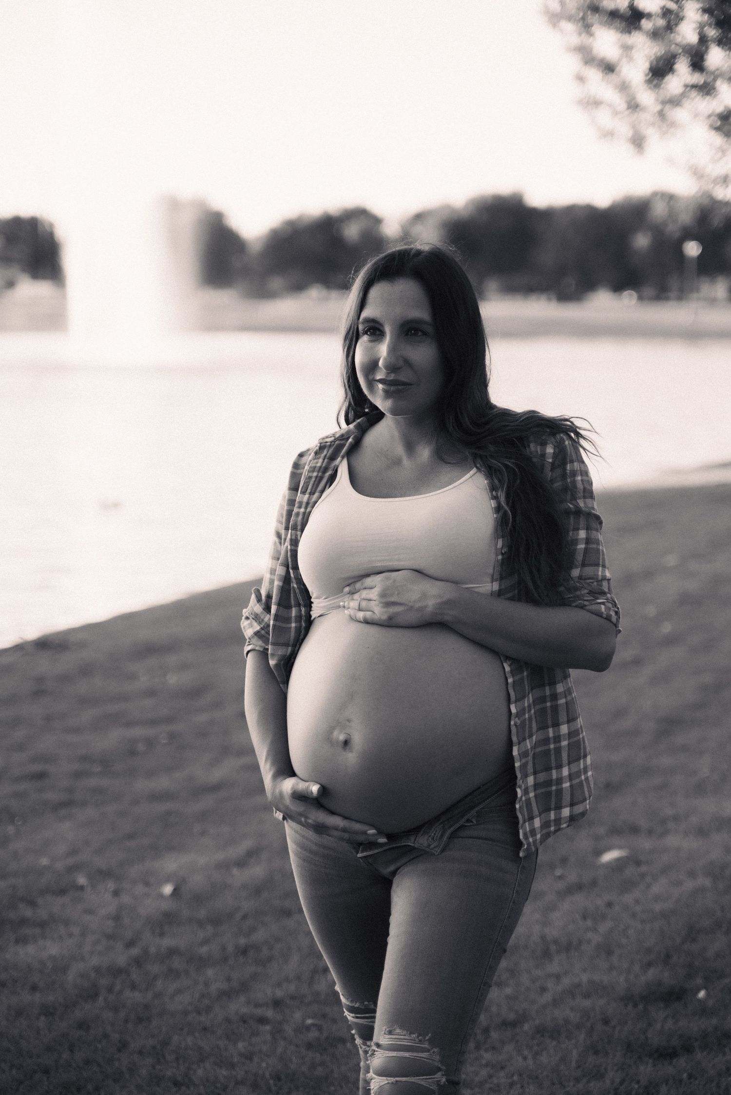 An artistic black and white portrait captures a pregnant woman in casual attire standing near a waterfront.