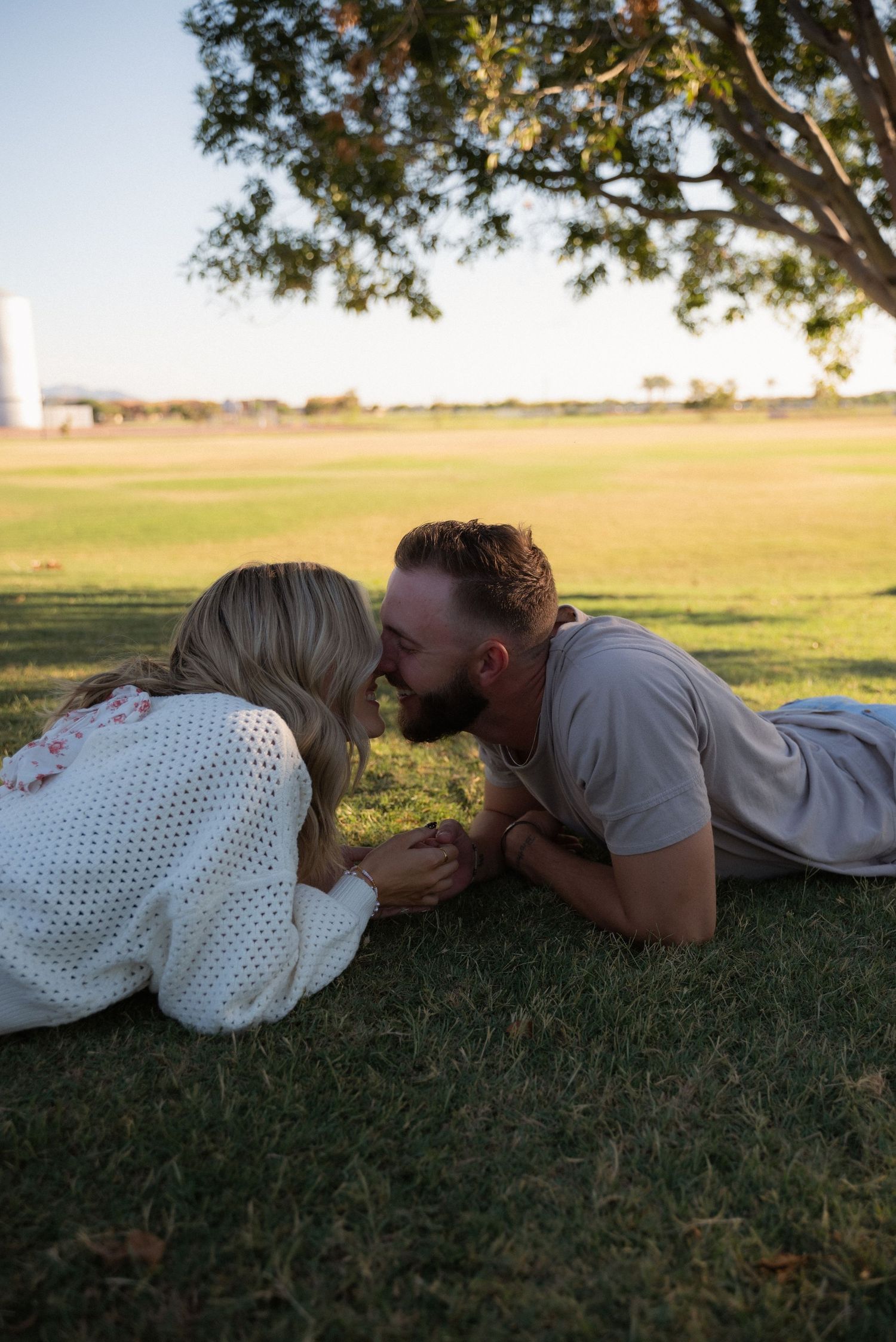 A romantic couple lying on grass in a park during sunset sharing an intimate moment.